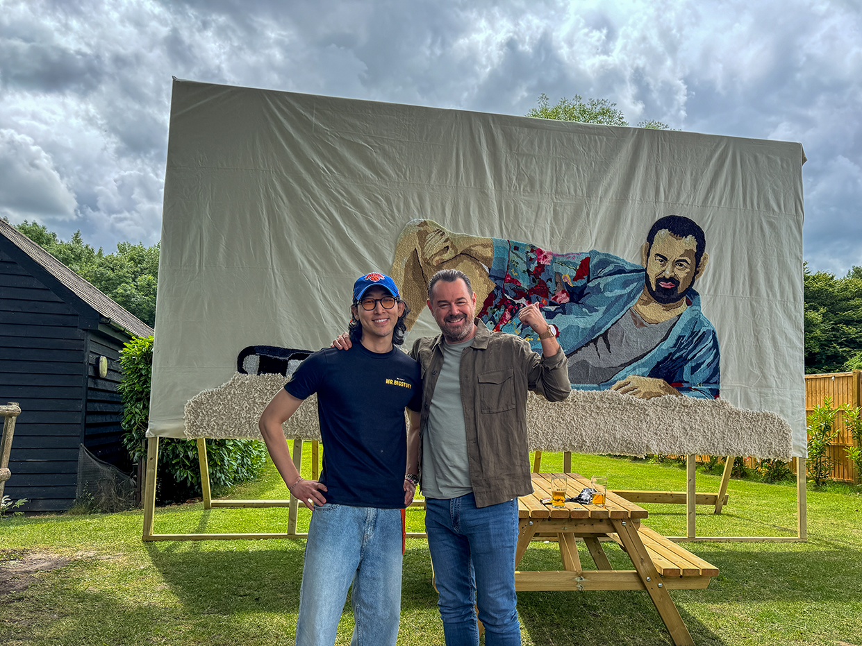 Two men standing arm in arm outdoors in front of a large framed textile artwork depicting a reclining man with a beard wearing a colorful robe.