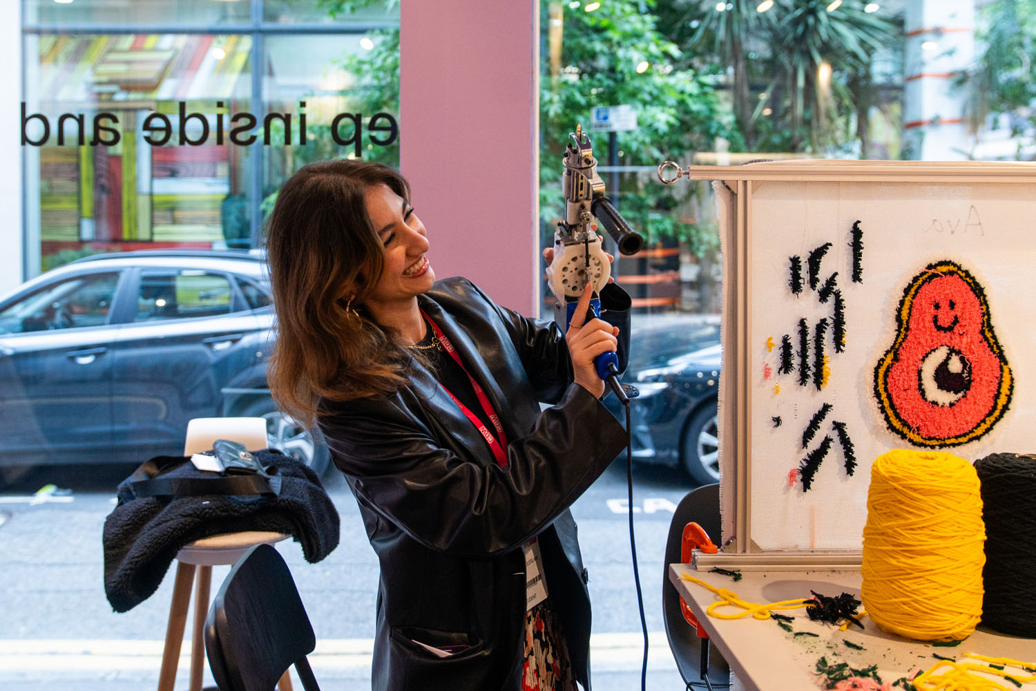 Smiling woman using a tufting gun to create colorful yarn art on a frame inside a workshop with a window showing cars outside.