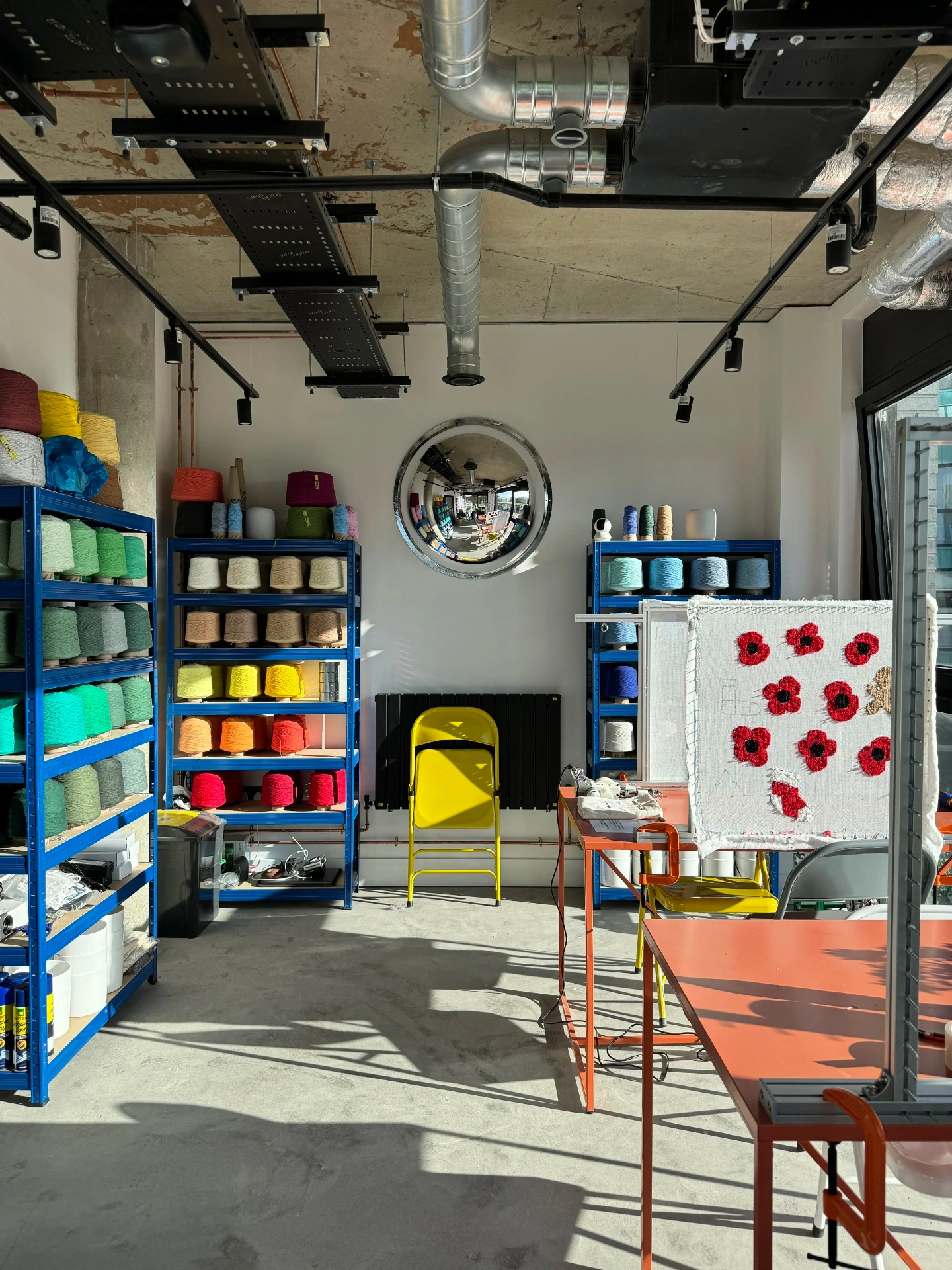 Bright craft room with blue shelves filled with colorful yarn skeins, a yellow chair, an embroidery frame with red poppy flowers, and exposed ceiling ducts.