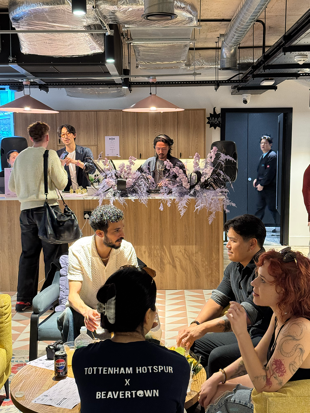 Group of people socializing in a modern indoor space with wooden counter decorated with purple flowers and a DJ playing music.
