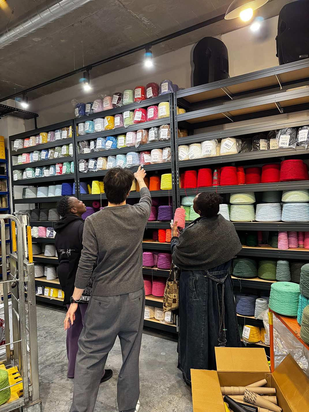Three people selecting colorful yarn from shelves in a yarn store or warehouse.