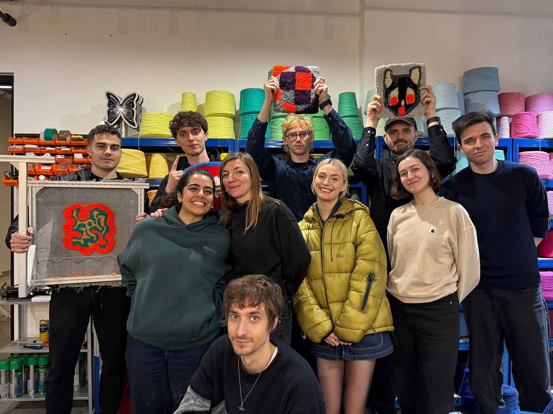Group of eleven people posing indoors with colorful yarn skeins on shelves behind them, some holding textile art pieces.