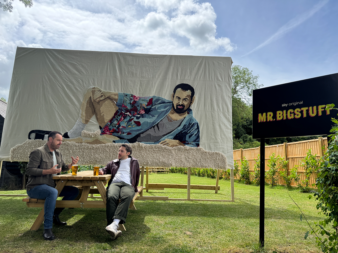 Two men sitting at a wooden picnic table with pints of beer, in front of a large textile artwork of a man reclining, with a sign nearby reading 'sky original MR. BIGSTUFF'.