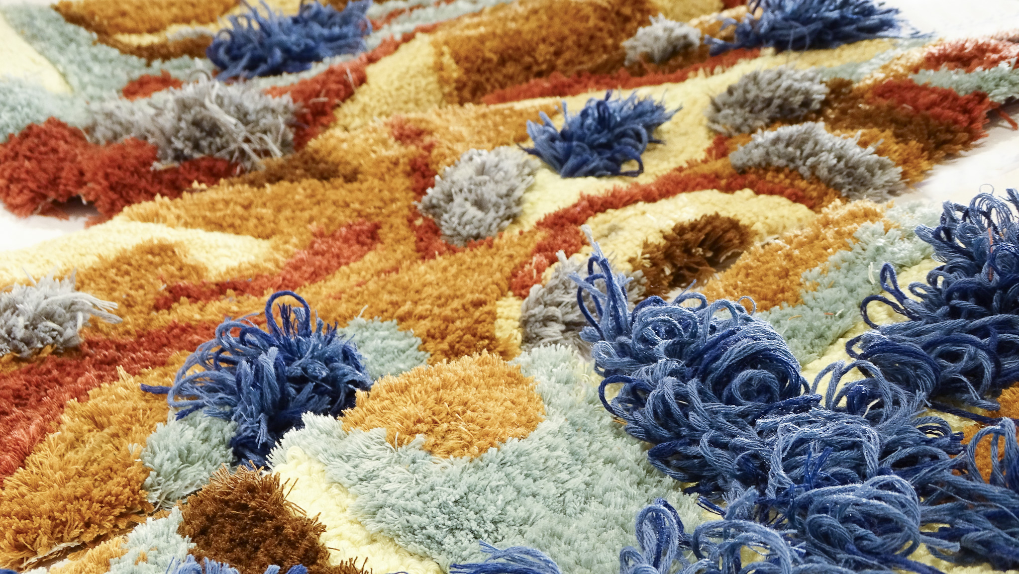 Close-up of a colorful textured fabric with tufted yarn clusters in blue, orange, yellow, and gray tones.