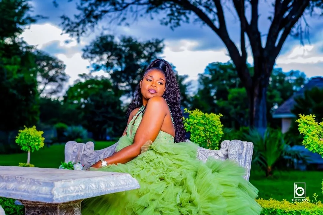 Woman in a voluminous green dress sitting on a decorative stone bench in a garden with trees and plants under a cloudy sky.