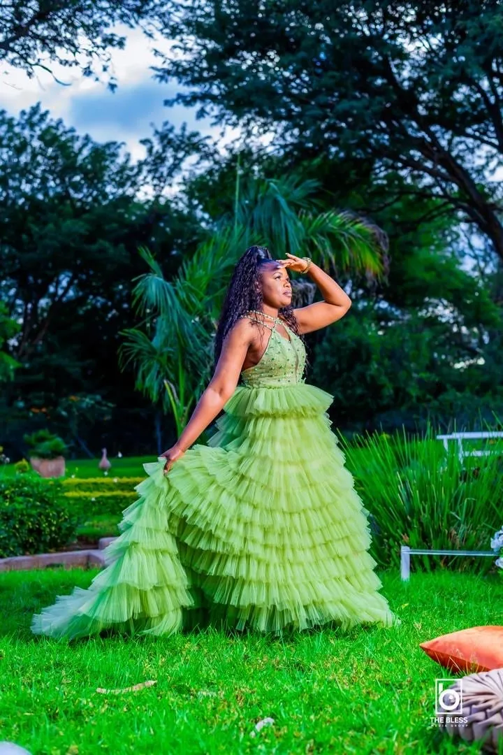 Woman in a green layered tulle gown standing on grass with her hand raised to her forehead, surrounded by trees and plants.