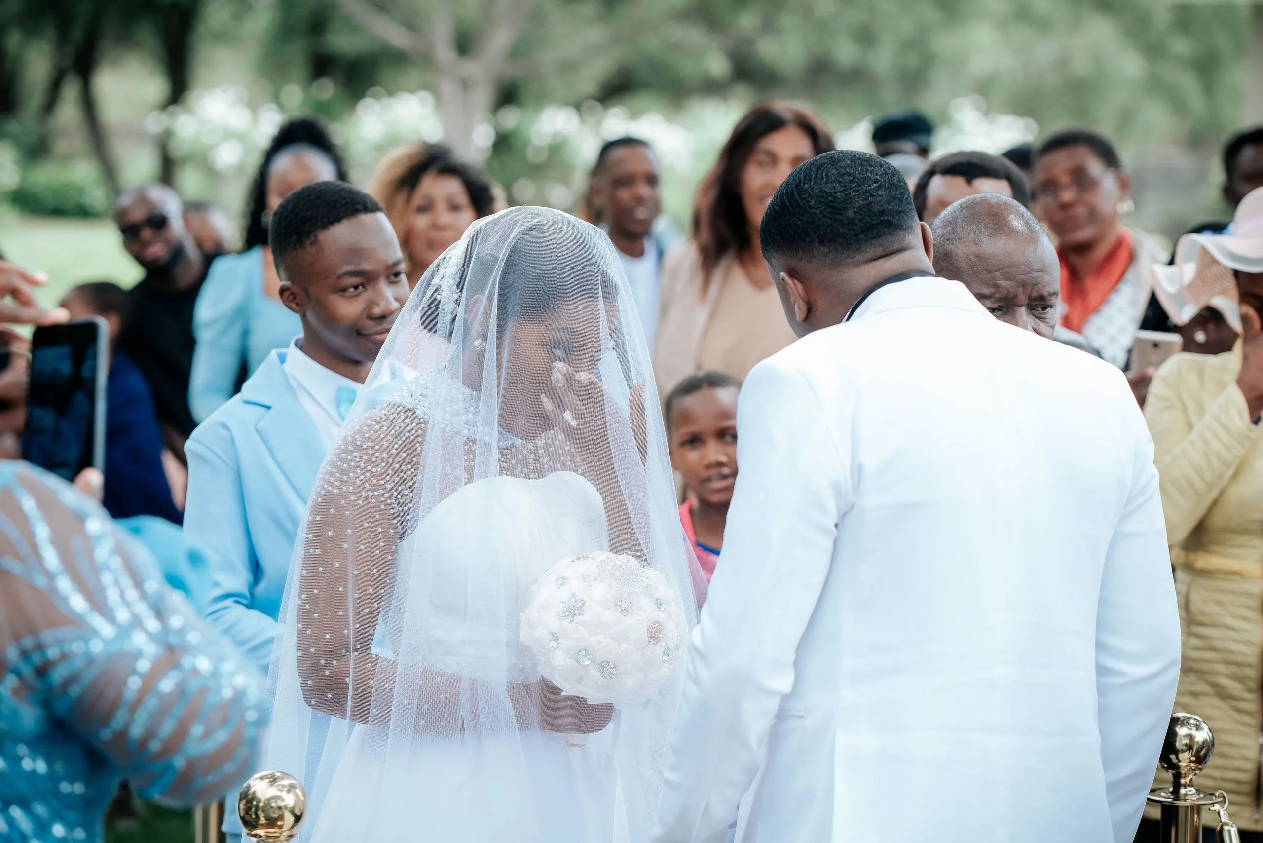 Bride in a white dress and veil wiping a tear, holding a bouquet, with groom in white suit and guests in the background at an outdoor wedding.