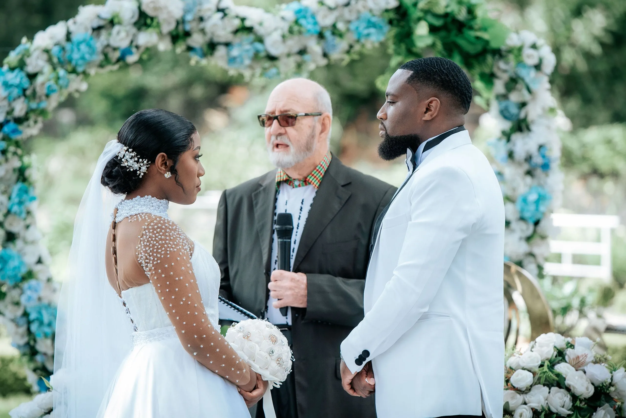 Bride and groom standing face to face during an outdoor wedding ceremony with the officiant holding a microphone between them under a floral arch.