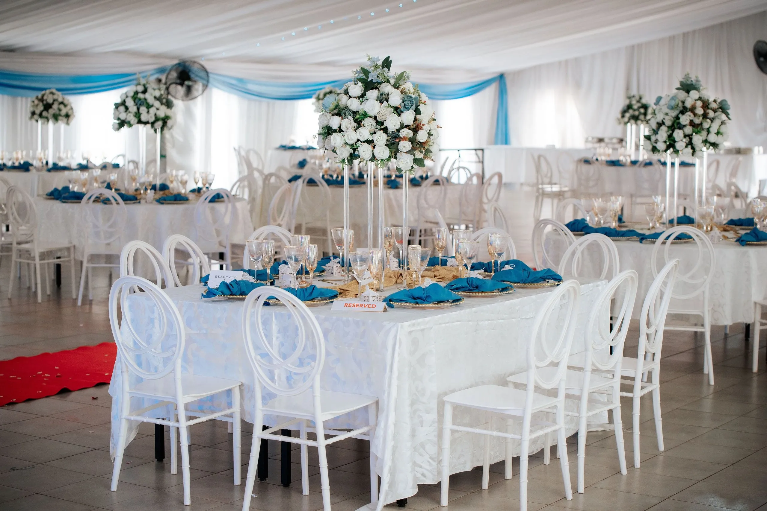 Elegant event hall with white tables set with blue napkins, glassware, gold plates, and tall floral centerpieces of white and green flowers.