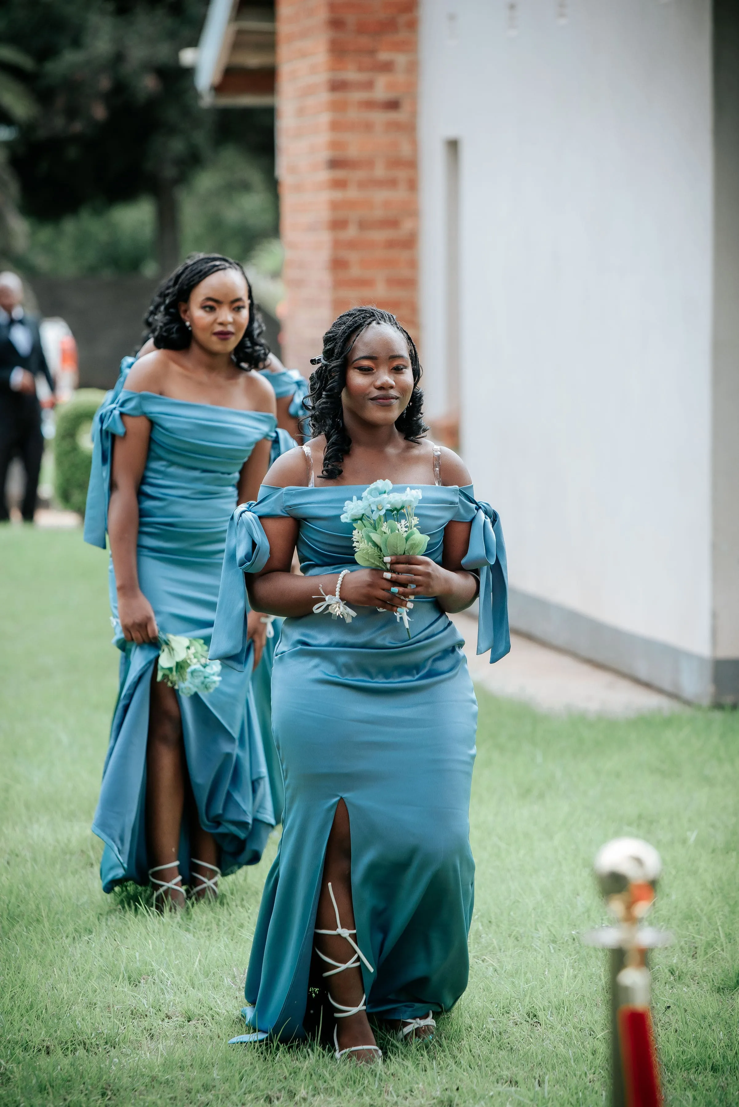 Two women in matching teal off-shoulder dresses walking outside on grass, each holding a small bouquet.