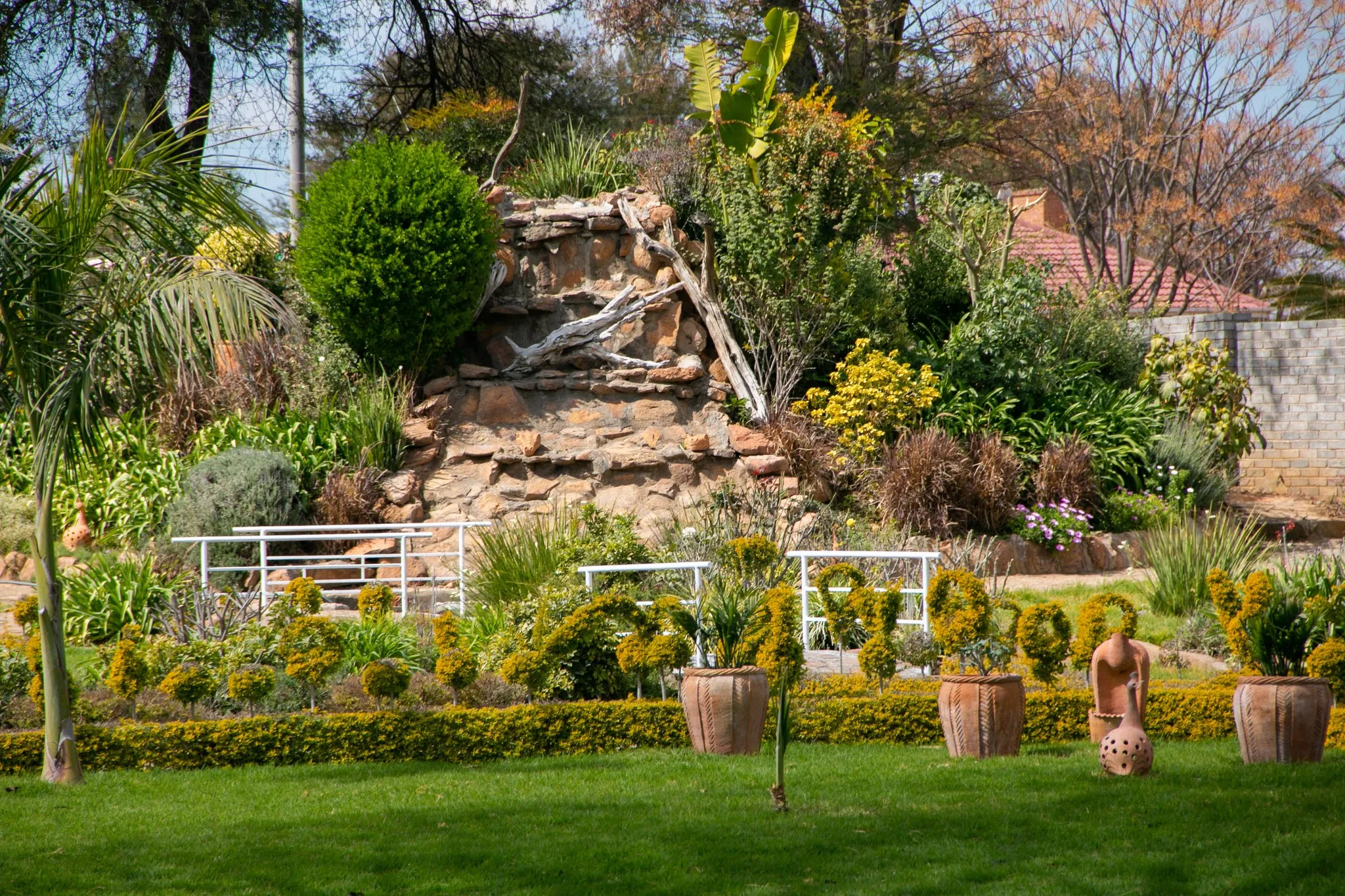Well-maintained garden with trimmed bushes, potted plants, green lawn, and a rock structure with a wooden branch.