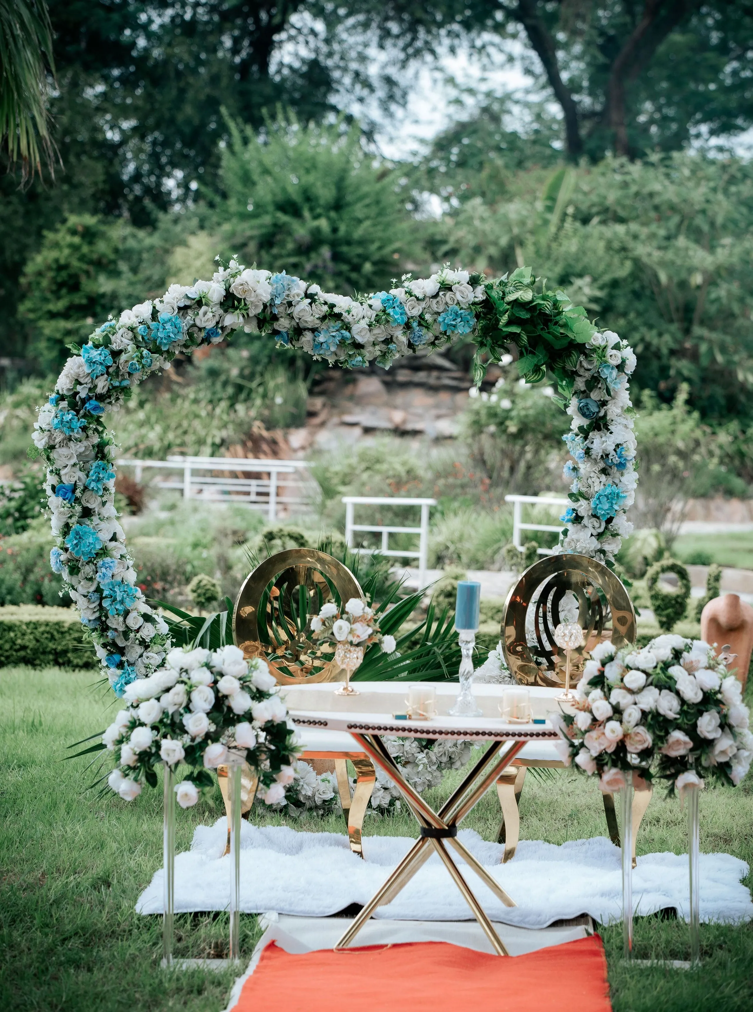 Outdoor wedding setup with a heart-shaped floral arch of white and blue flowers, gold chairs, and a table with floral arrangements and candles.