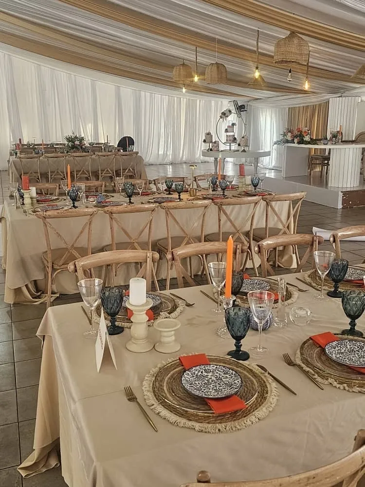 Indoor event setup with long tables covered in beige cloth, wooden chairs, and table settings including blue patterned plates, orange napkins, candles, and elegant glassware under striped ceiling drapes and wicker hanging lights.