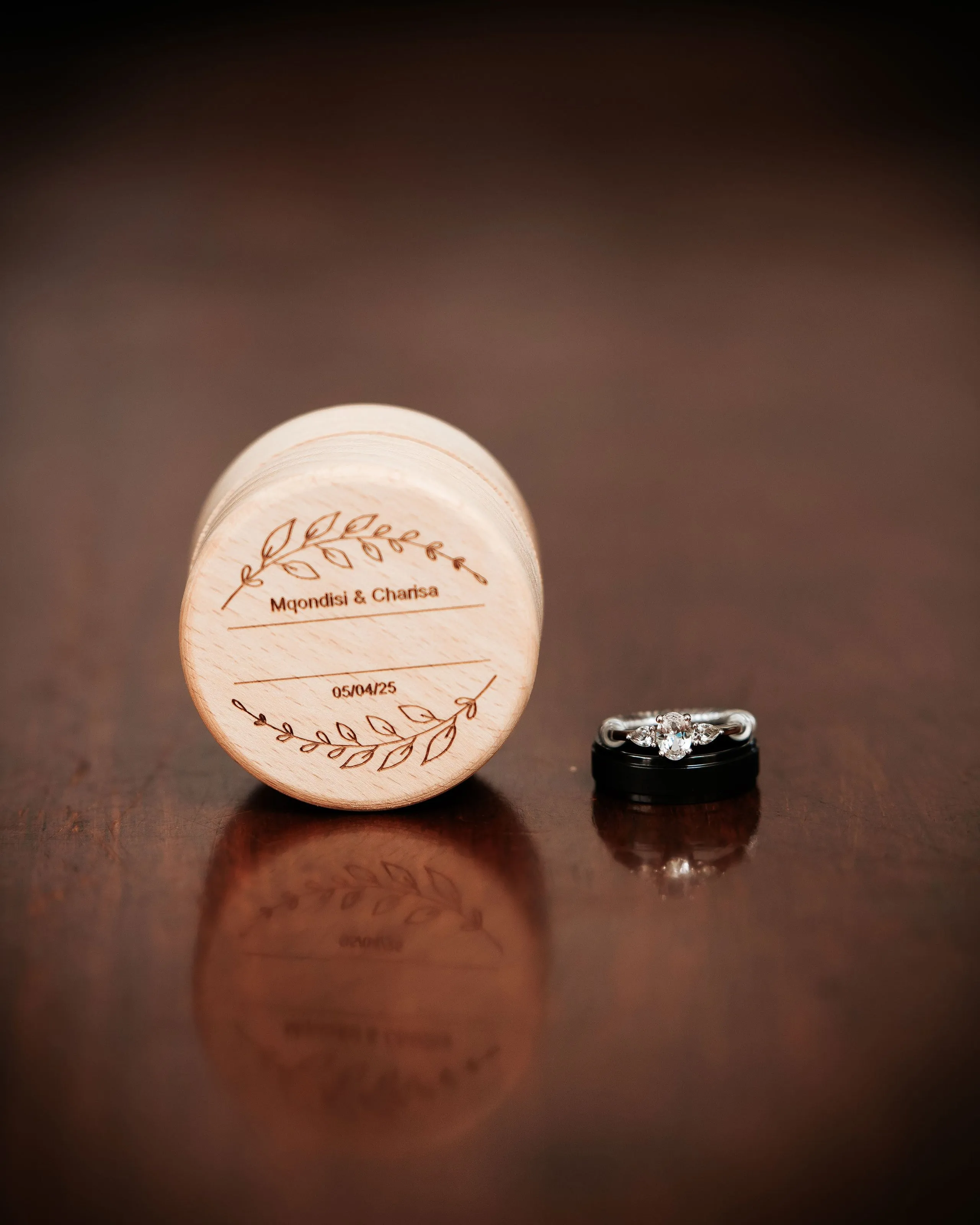 Wooden circular box with engraved leaves and text 'Mqondisi & Charisa' and date '05/04/25' beside a set of wedding rings on a wooden surface.