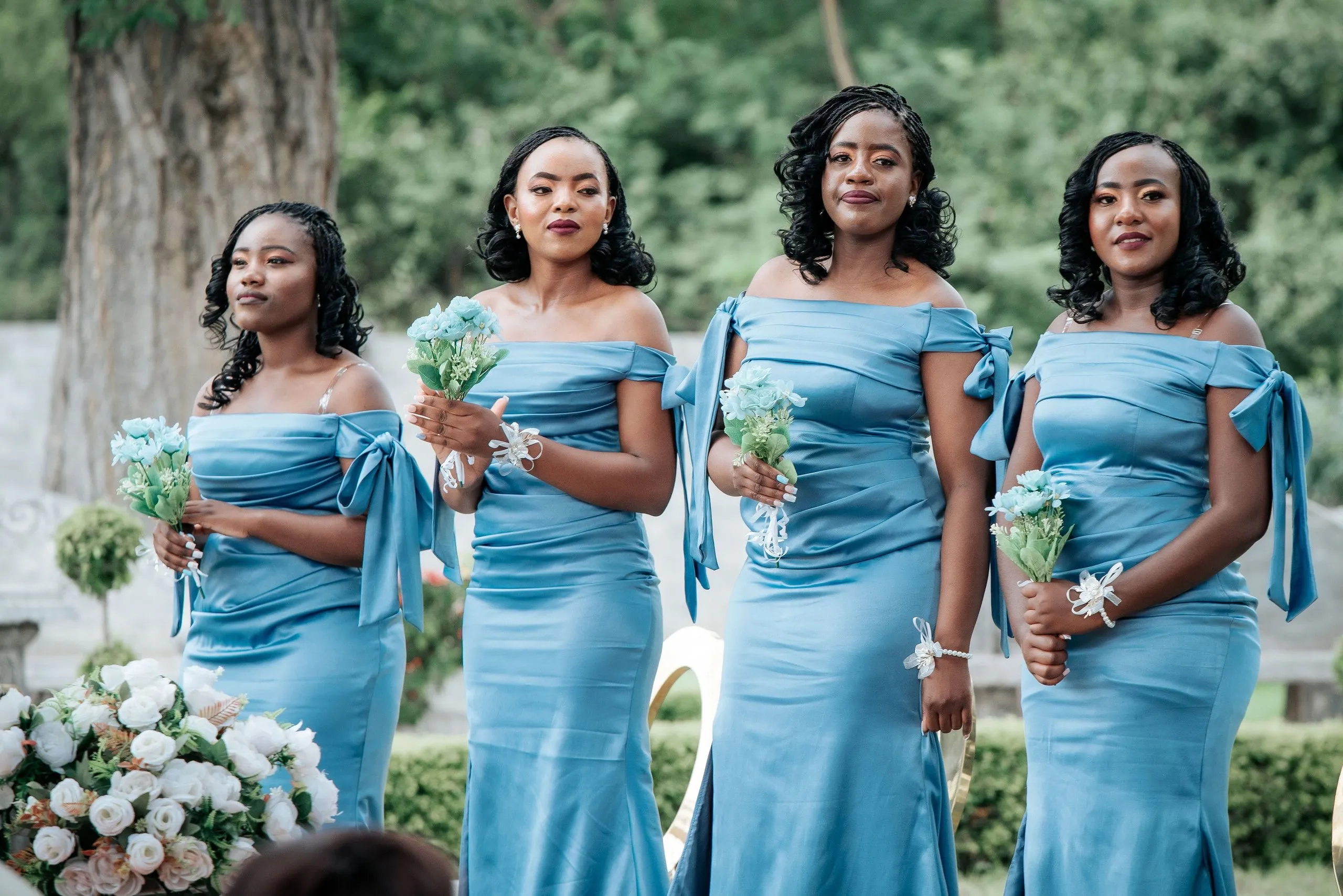 Four bridesmaids standing outdoors in matching blue off-shoulder dresses holding blue flower bouquets.