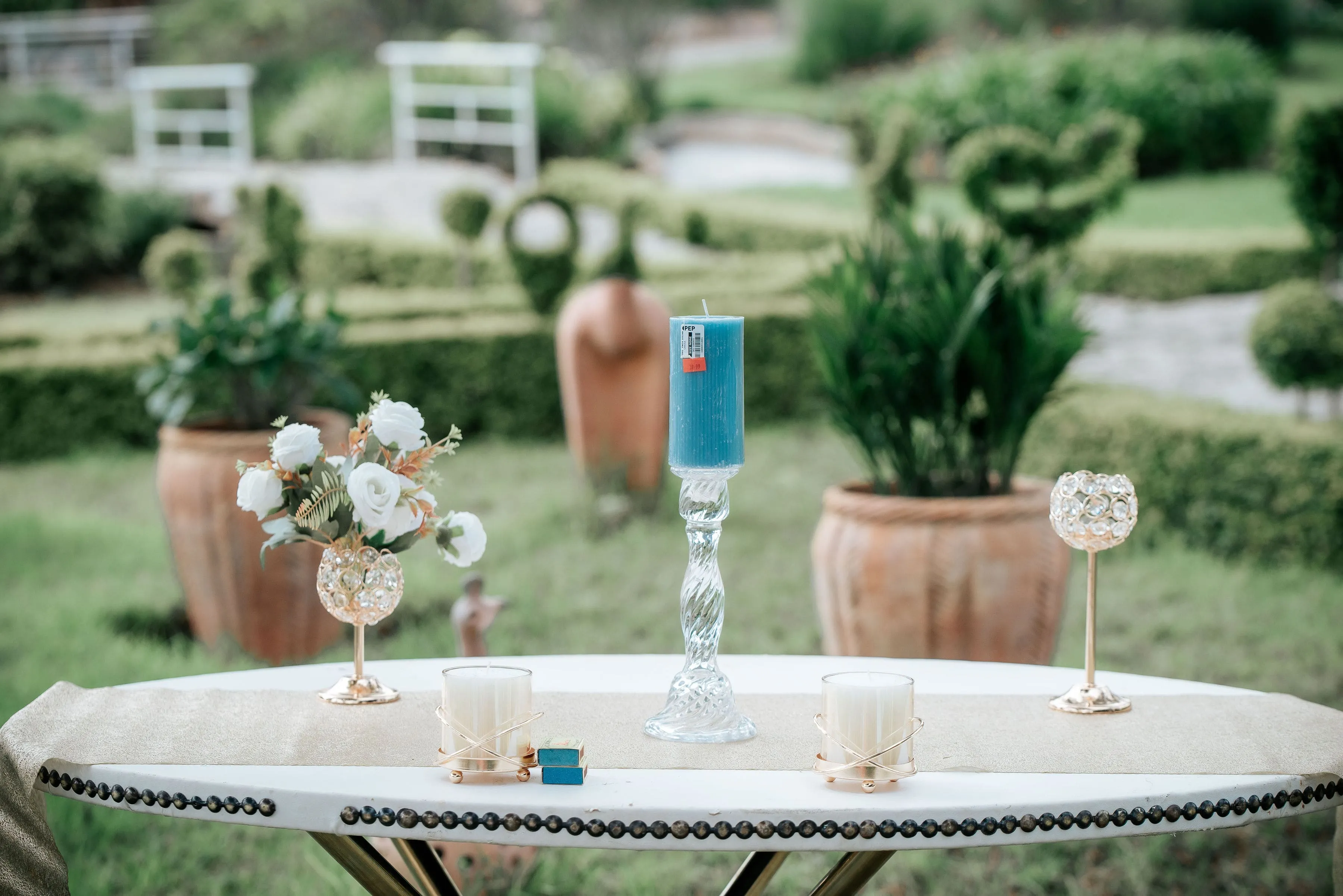 Outdoor table set with a tall blue candle on a glass holder, two white votive candles in gold holders, a small bouquet of white flowers in a gold vase, and two matchboxes, with green garden background.