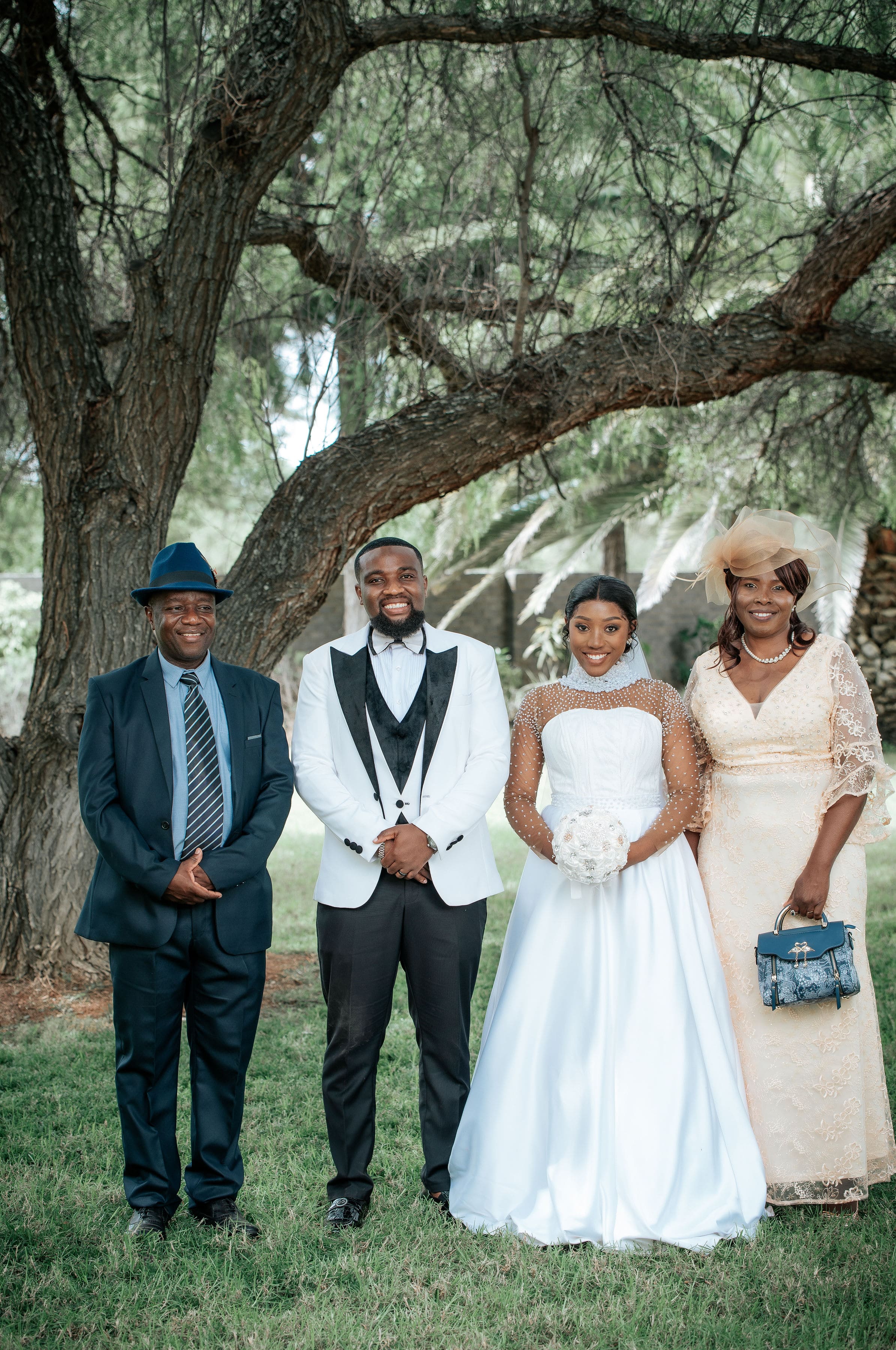 Bride in white gown holding a bouquet and groom in white tuxedo standing with two adults under a large tree.