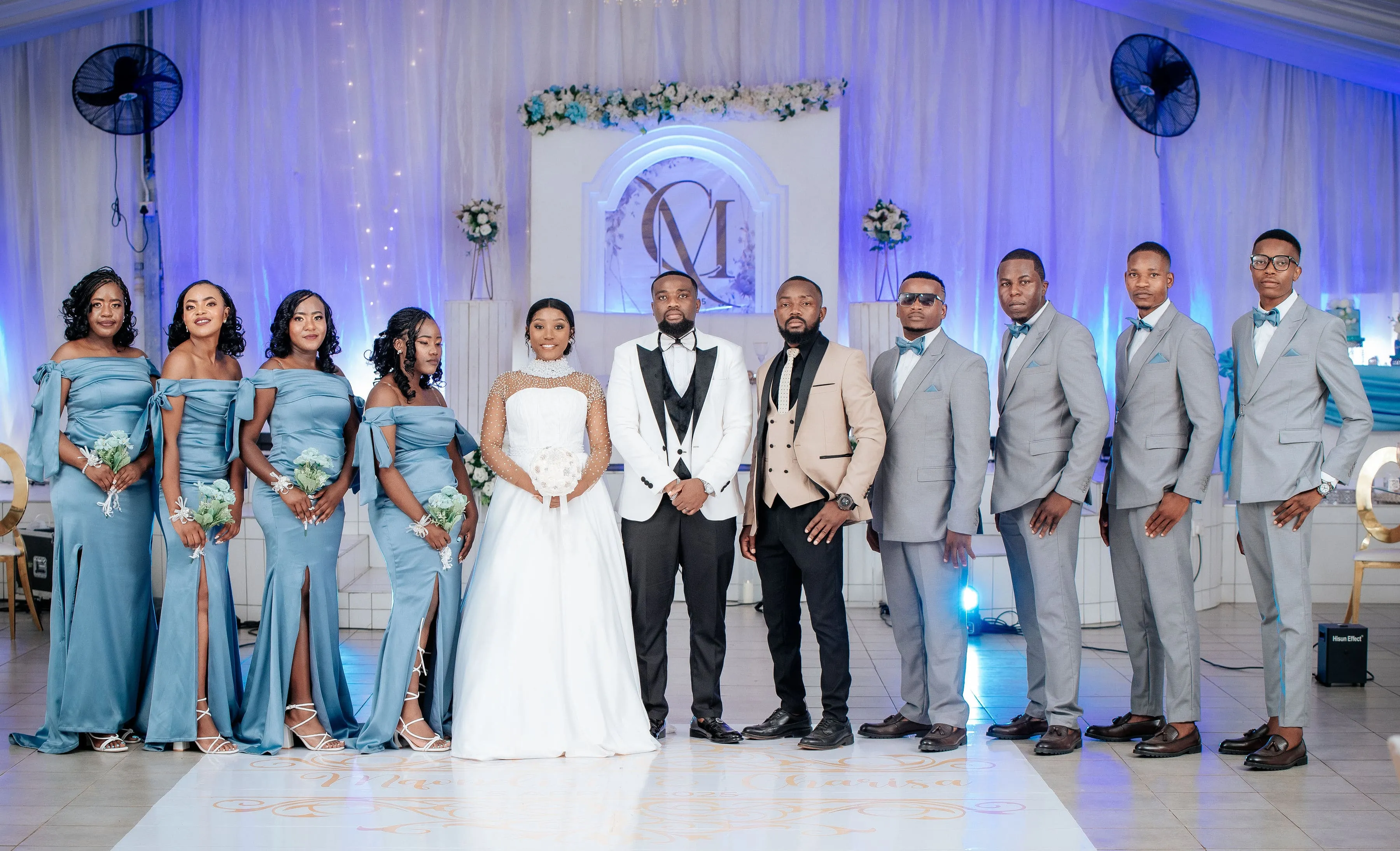 Bride in white gown and groom in white tuxedo with bridal party in blue dresses and gray suits standing indoors against a white and blue floral decorated backdrop.