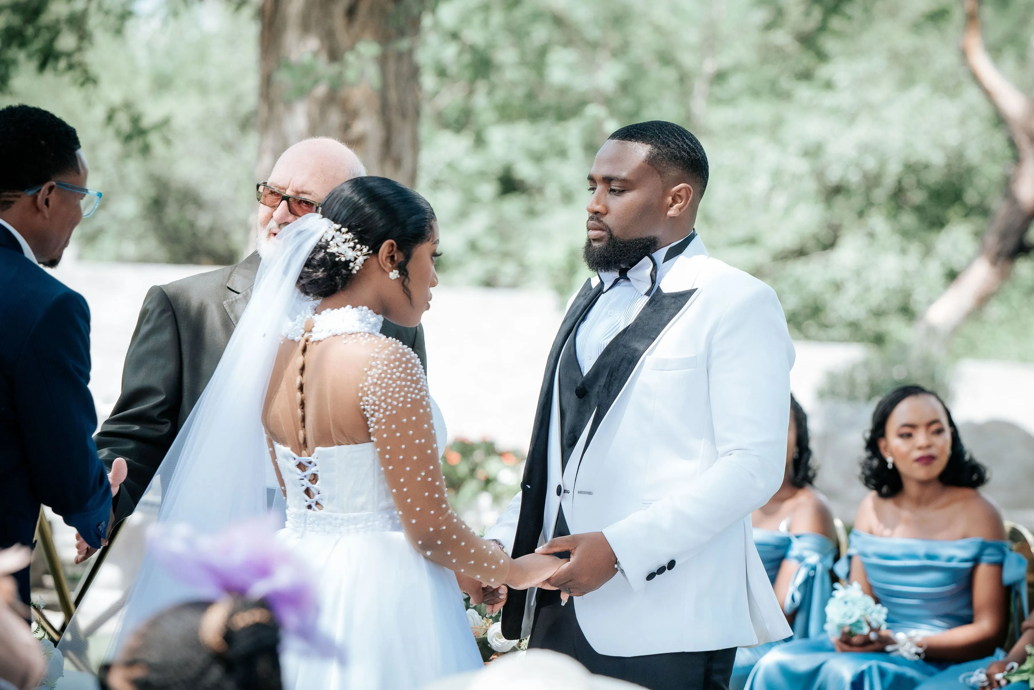 Bride and groom holding hands during an outdoor wedding ceremony with guests in blue dresses in the background.