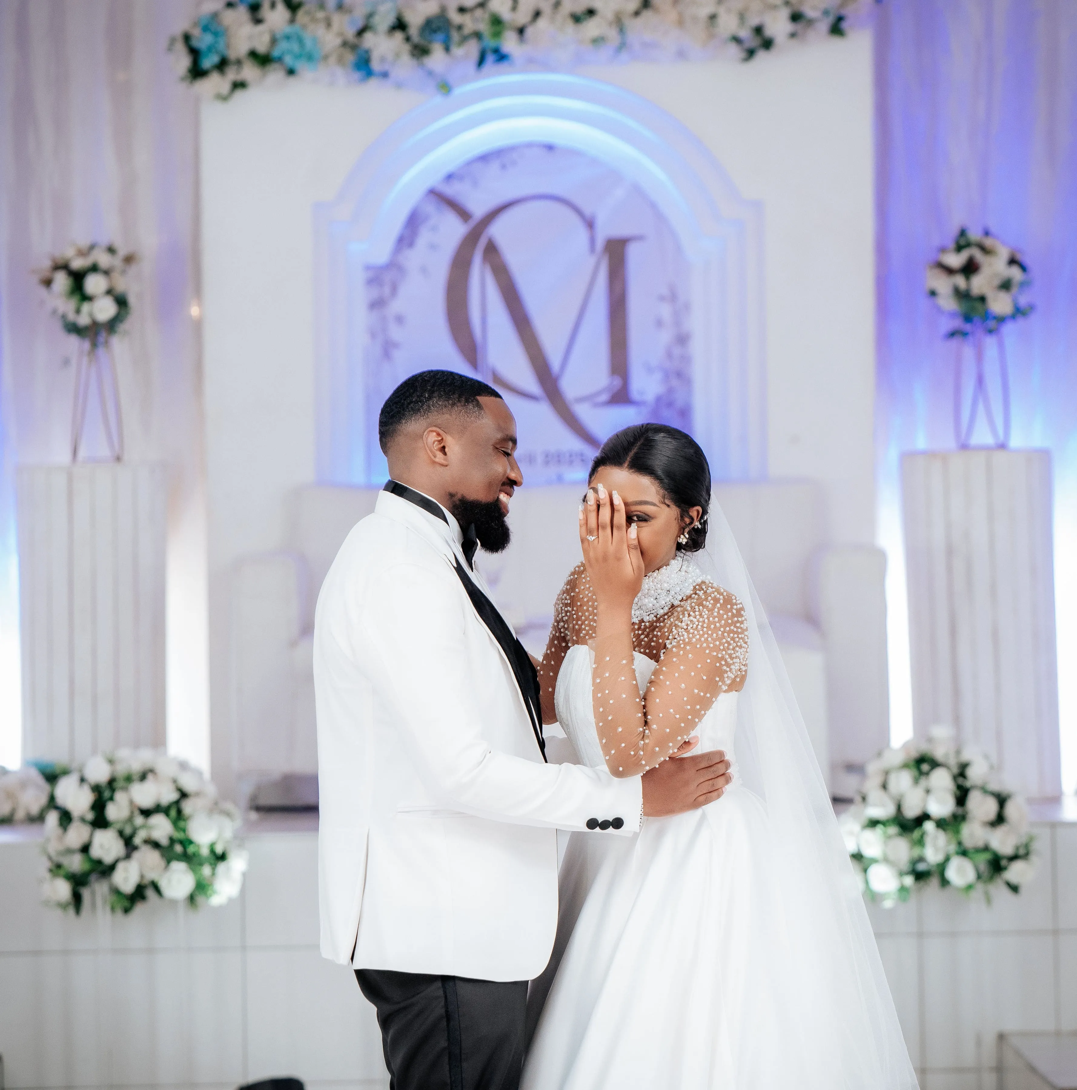 Bride in white dress covering part of her face with her hand while groom in white jacket embraces her during wedding ceremony.