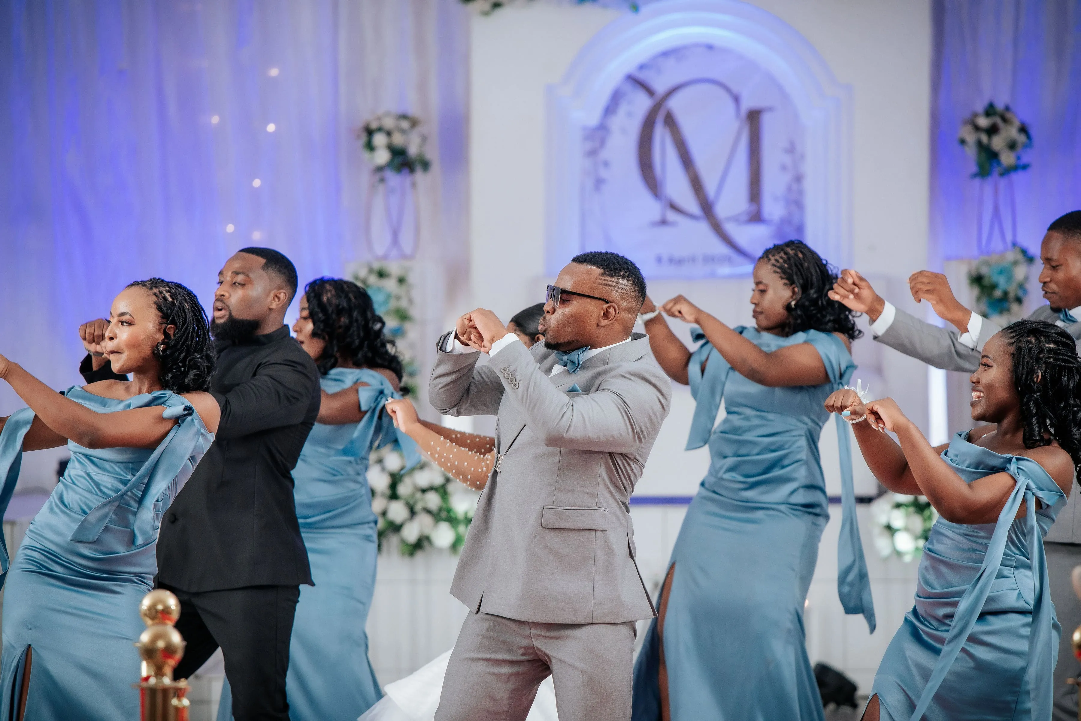 Group of people dancing indoors at a formal event, with women in blue dresses and men in suits.