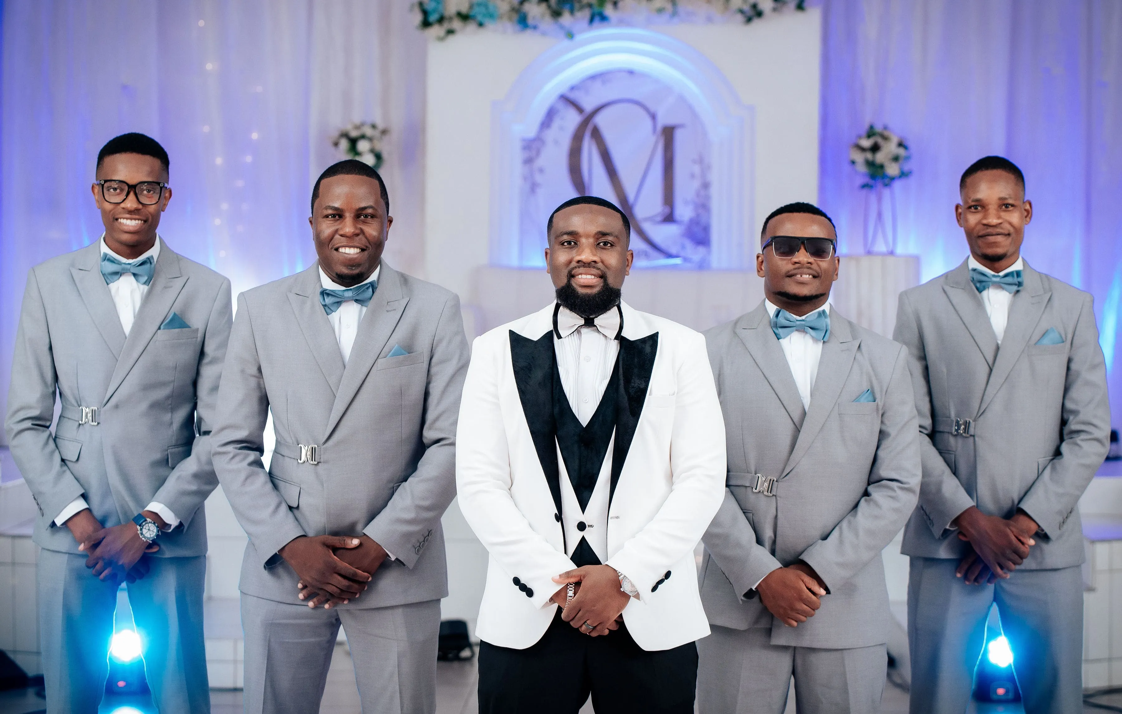 Group portrait of five men at a formal event with one man in a white tuxedo jacket and four men in matching gray suits with blue bow ties.