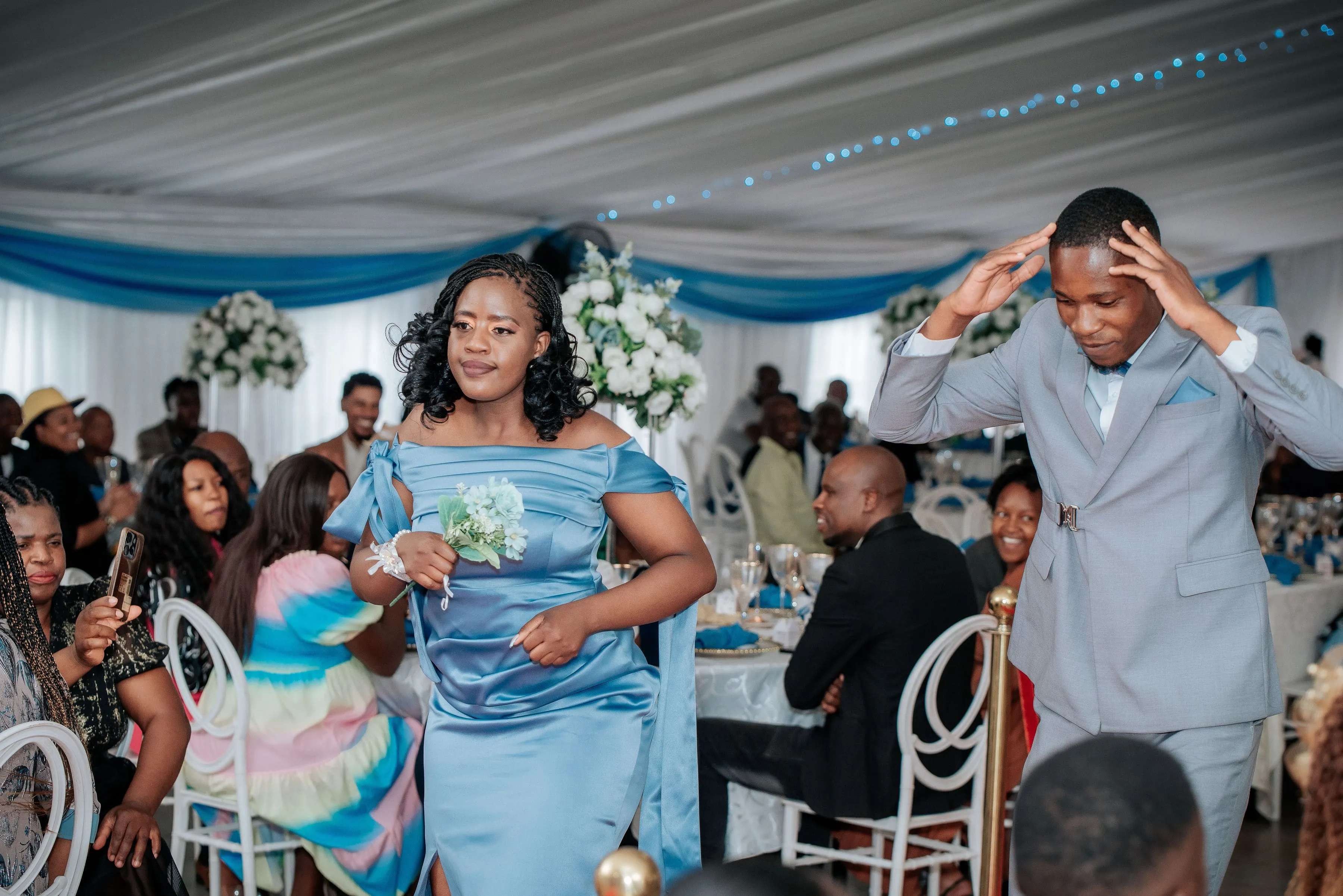 Woman in a blue satin dress holding a small bouquet walking beside a man in a light gray suit gesturing with his hands at a decorated indoor event with seated guests.