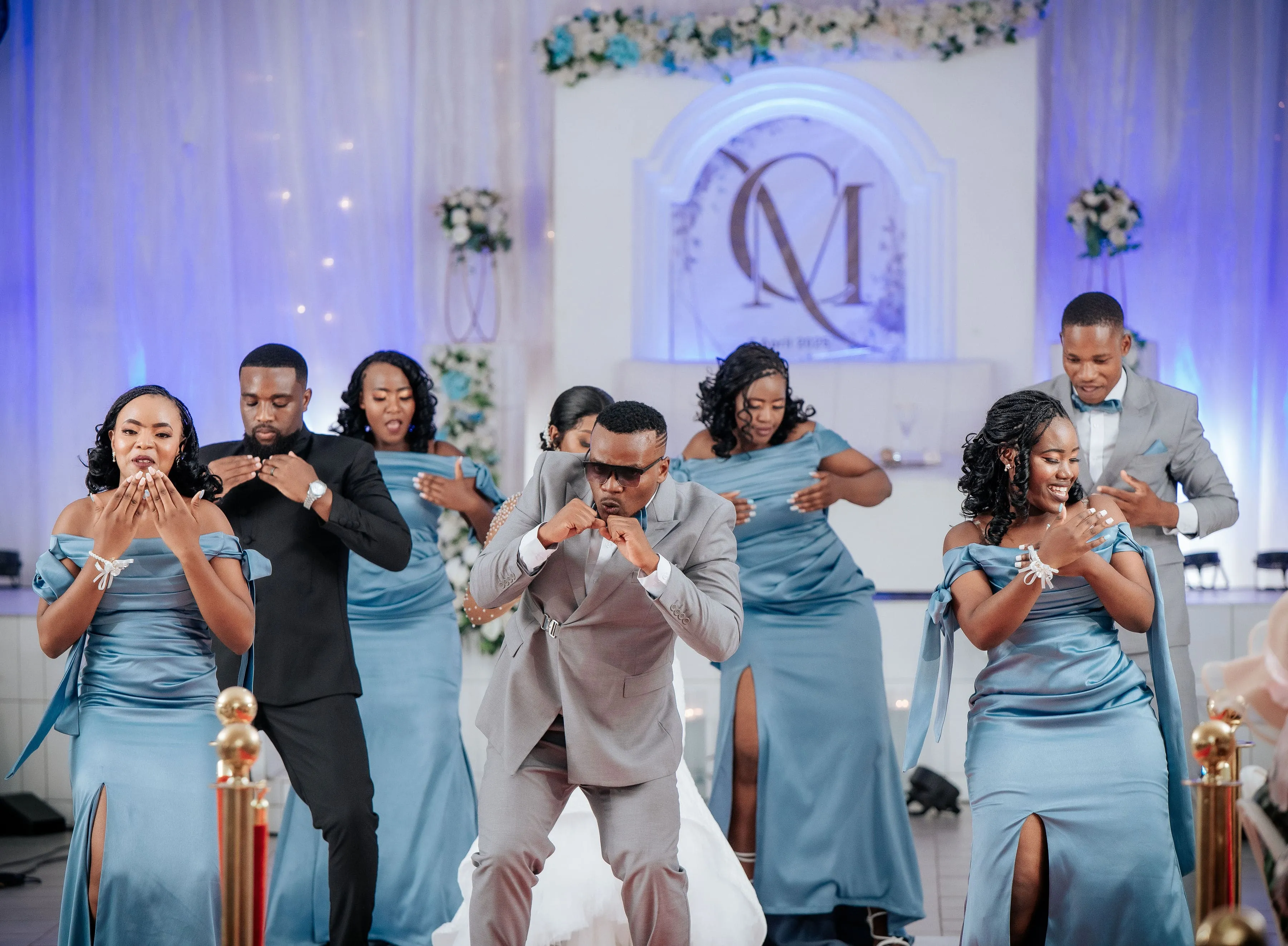 Group of bridesmaids in light blue dresses and groomsmen in suits performing a coordinated dance at a wedding reception.