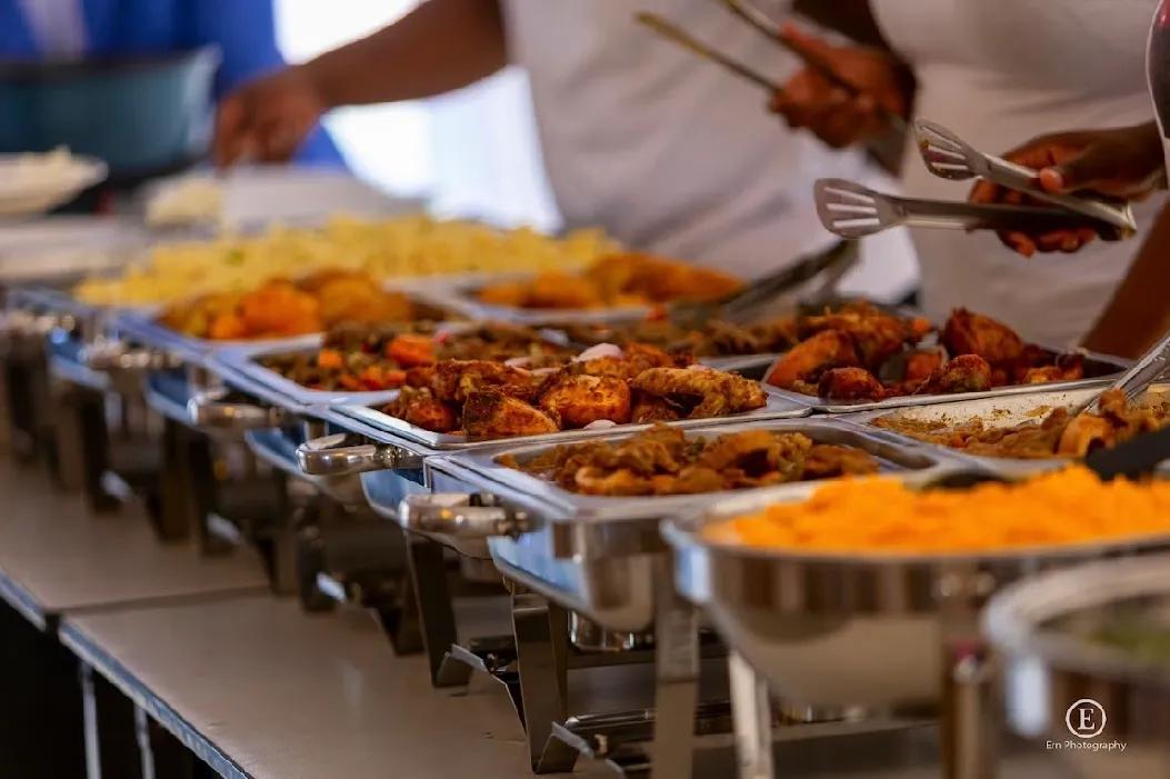 Buffet table with trays of assorted cooked foods including rice and meat dishes, with hands serving using tongs.