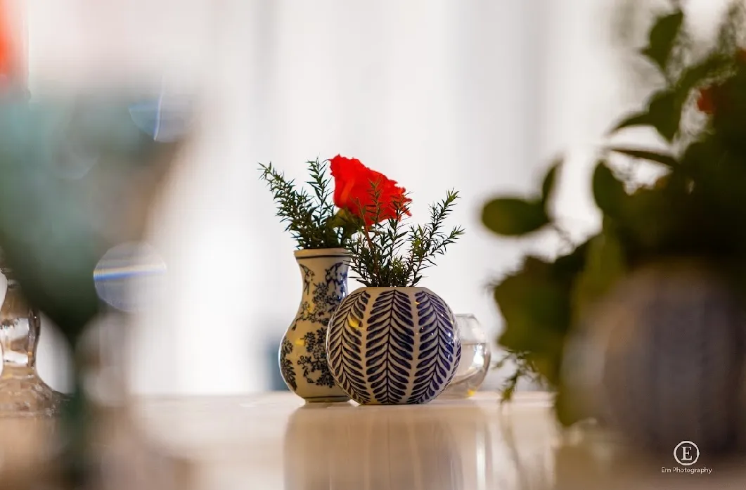 Two blue and white ceramic vases on a table, one round and one hourglass-shaped, with greenery and a single red flower in the hourglass vase.