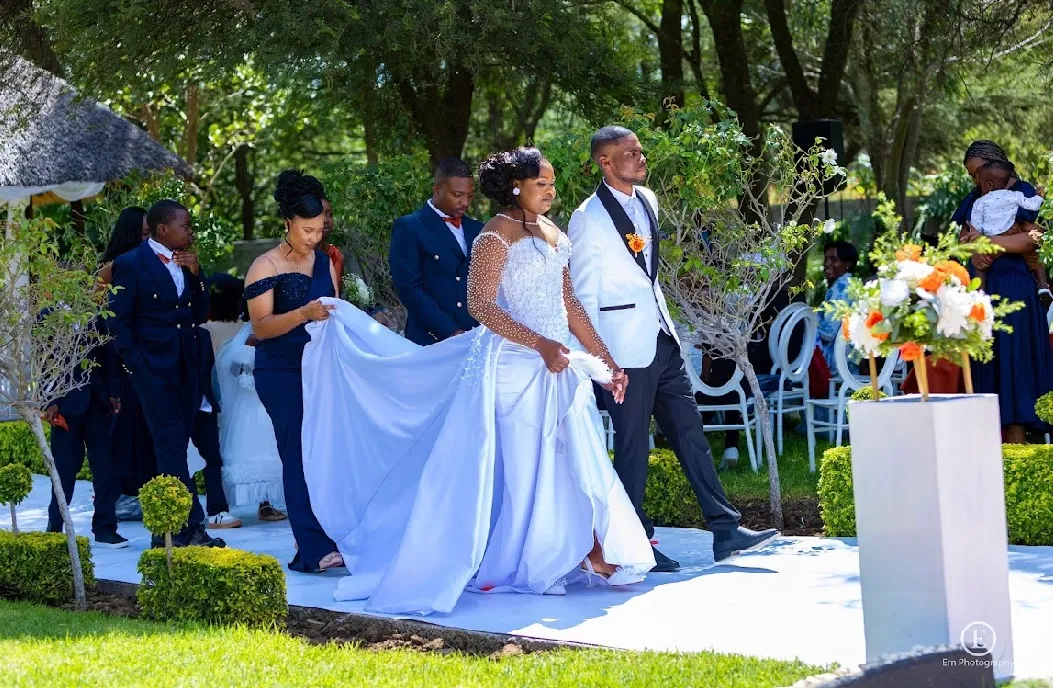Bride in a white wedding dress and groom in white tuxedo jacket walking down an outdoor aisle accompanied by attendants in formal attire.