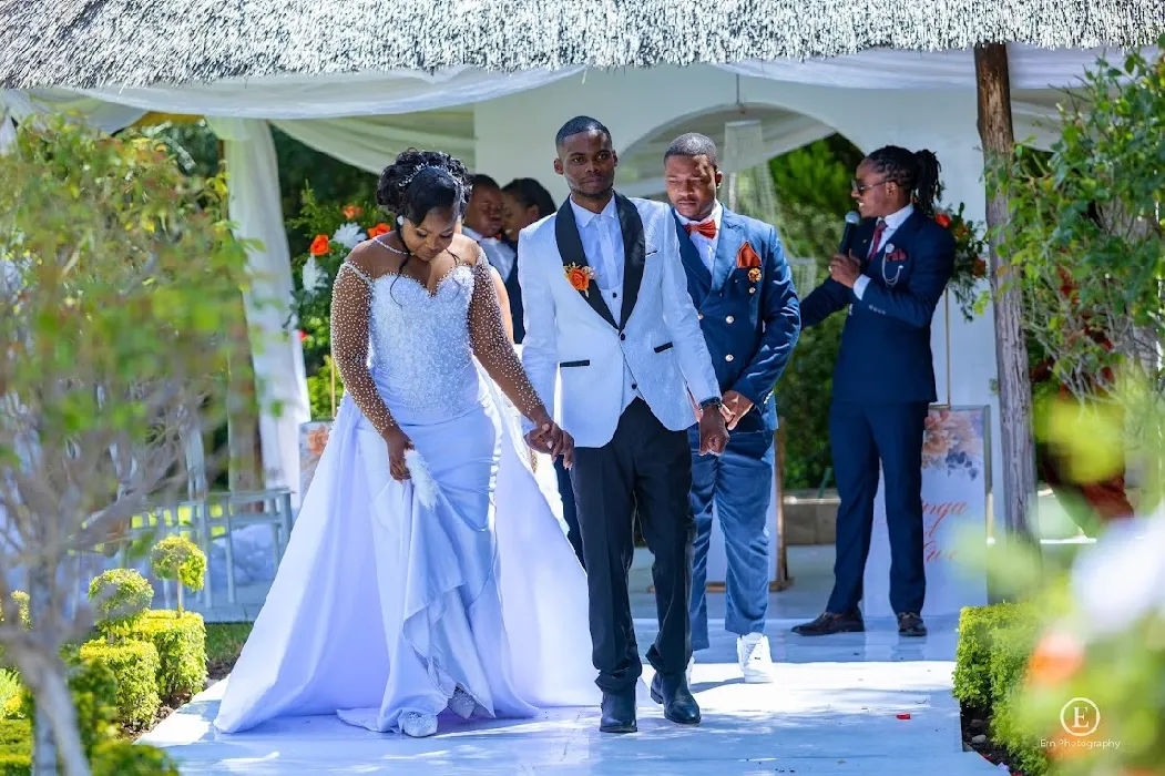 Bride and groom holding hands walking down an outdoor aisle with another man and an officiant speaking into a microphone in the background.