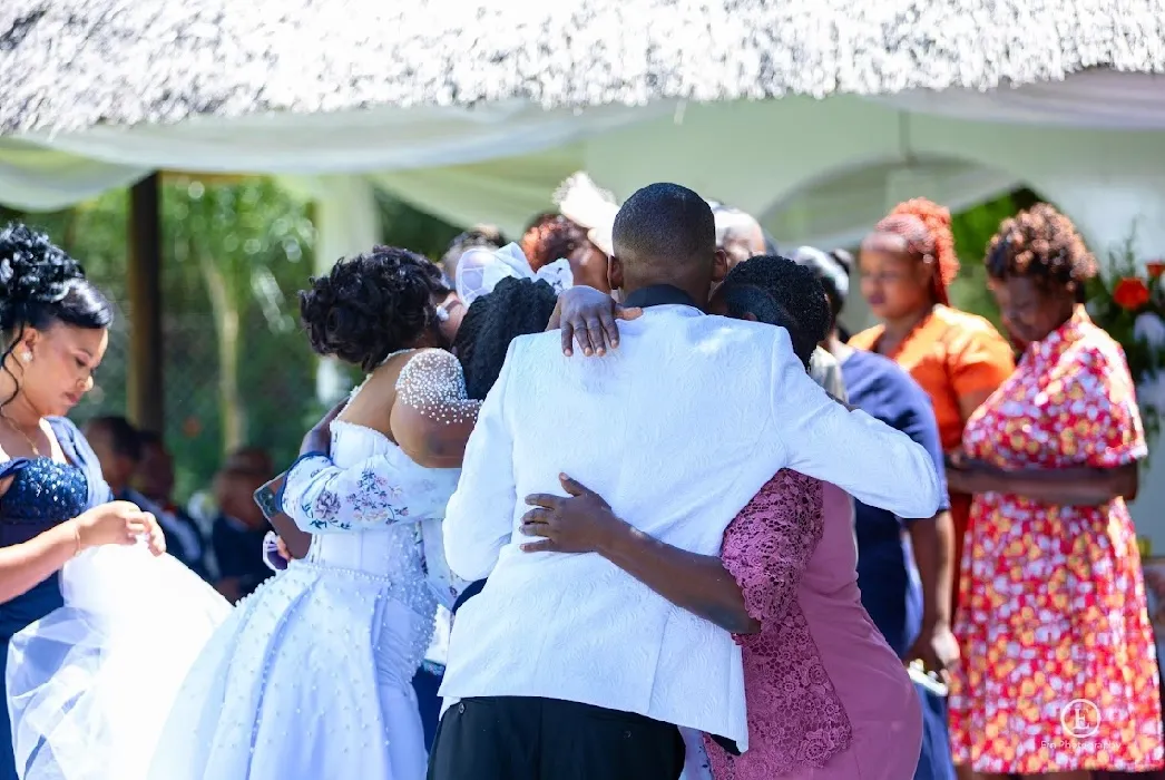 Group of people embracing at an outdoor event, with a man in a white suit jacket at the center.