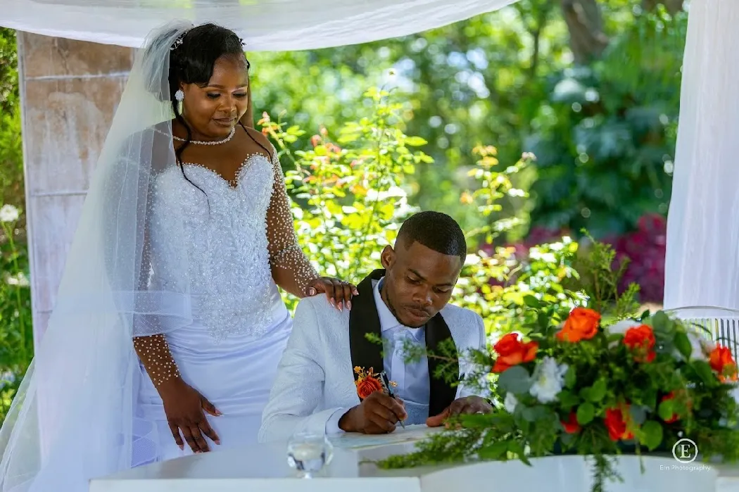 Bride in white wedding dress with veil standing beside groom in white tuxedo signing a document at a decorated outdoor table.