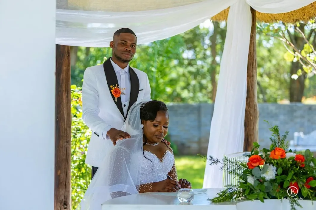Bride signing a document at a table with groom standing behind her, outdoors under a white canopy with greenery in the background.
