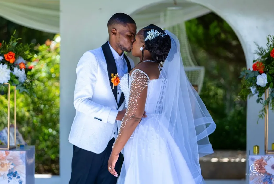 Bride and groom in wedding attire sharing a kiss under a white arch decorated with flowers.