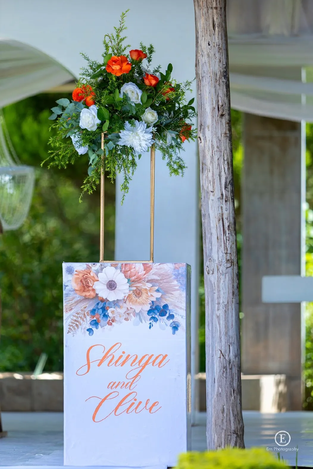 White sign with floral decoration and orange script reading 'Shinga and Clive' topped with an arrangement of orange and white flowers with green foliage.