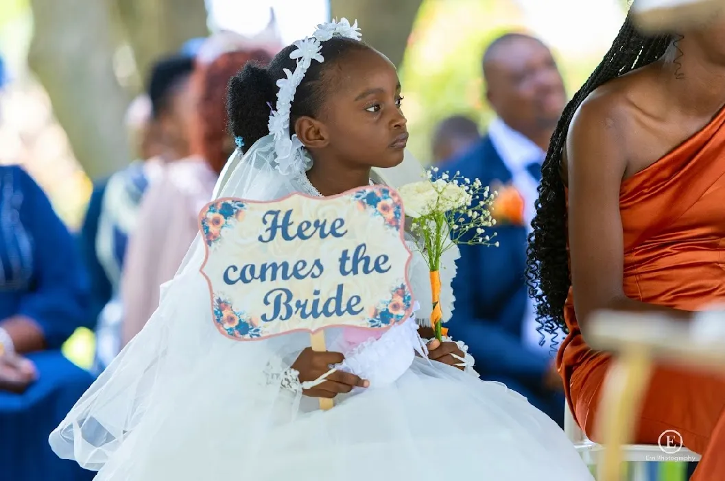 Young flower girl in white dress and veil holding a bouquet and a sign that says 'Here comes the Bride' at a wedding ceremony.