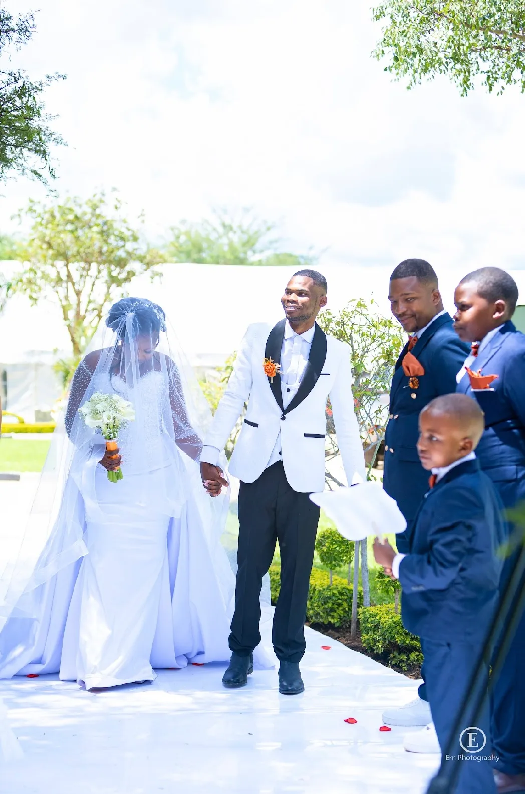 Bride in white wedding dress and veil holding a bouquet and groom in white tuxedo jacket holding hands outside with groomsmen in blue suits.
