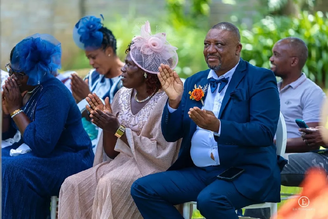 A group of well-dressed people sitting in a row outdoors, clapping and smiling, with women wearing decorative hats and a man in a blue suit with an orange boutonniere.