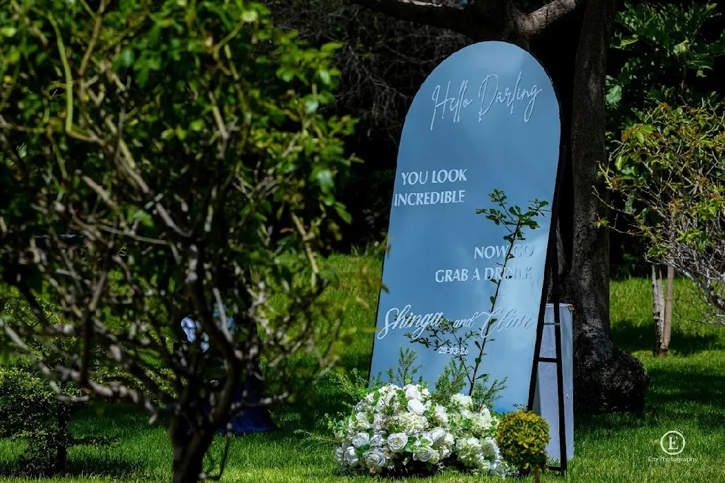 Blue wedding signboard with white text reading 'Hello Darling, You look incredible, Now go grab a drink, Shingai and Claire, 28.03.20', placed on grass with flower arrangement below and trees around.
