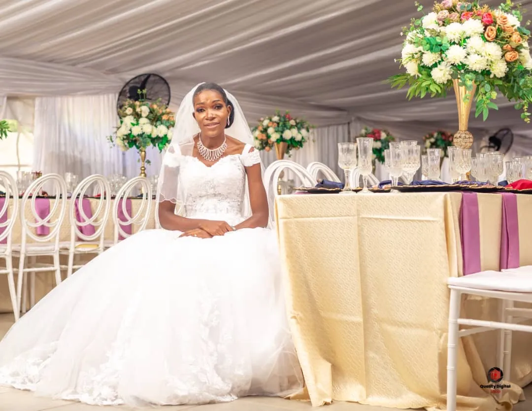 Bride in a white wedding dress sitting next to a decorated table with floral arrangements and glasses inside a tent.