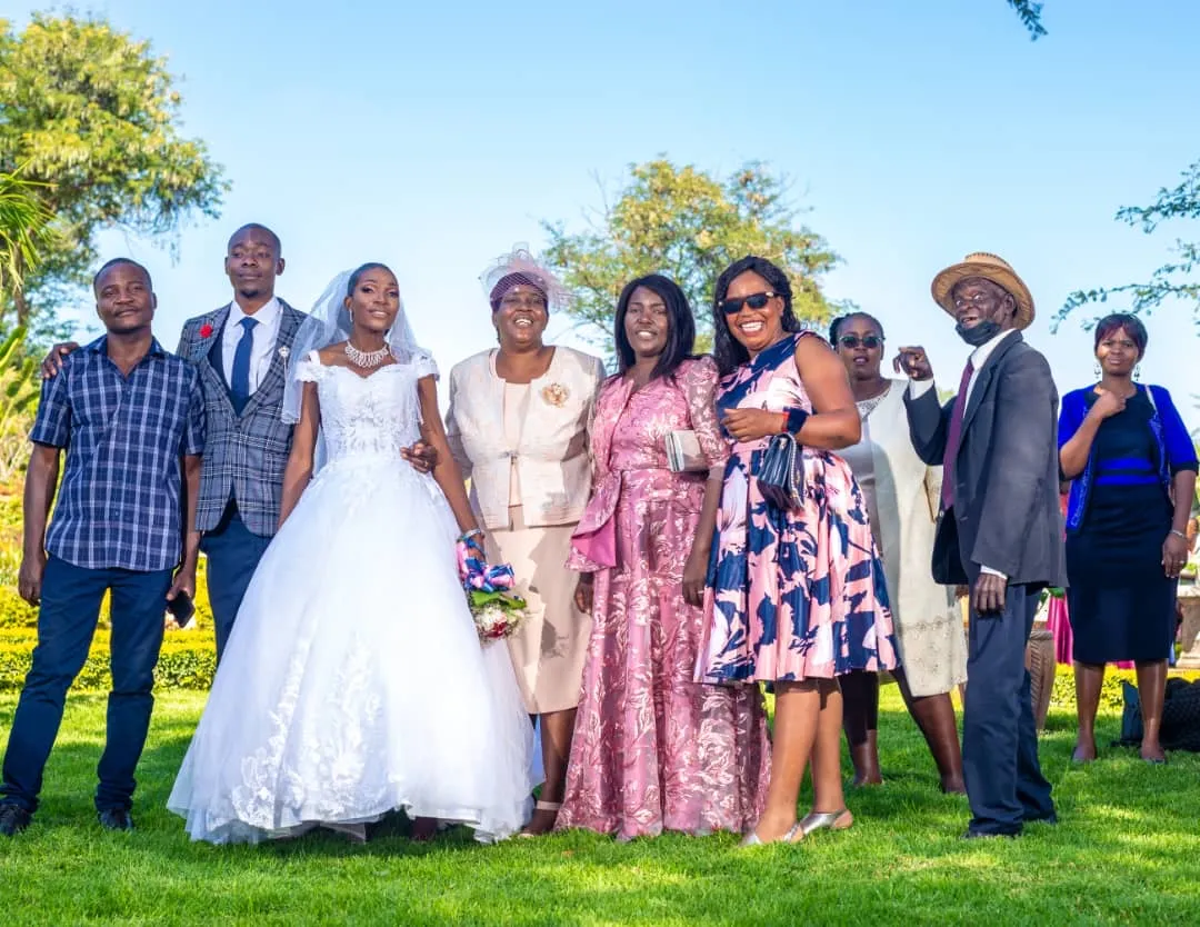 Bride in white wedding gown and groom in suit posing with smiling family and friends outdoors on green grass.