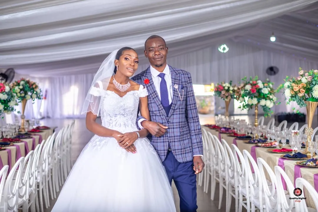 Bride in white wedding gown and veil linked arm-in-arm with groom in blue plaid suit and tie inside a flower-decorated reception hall.