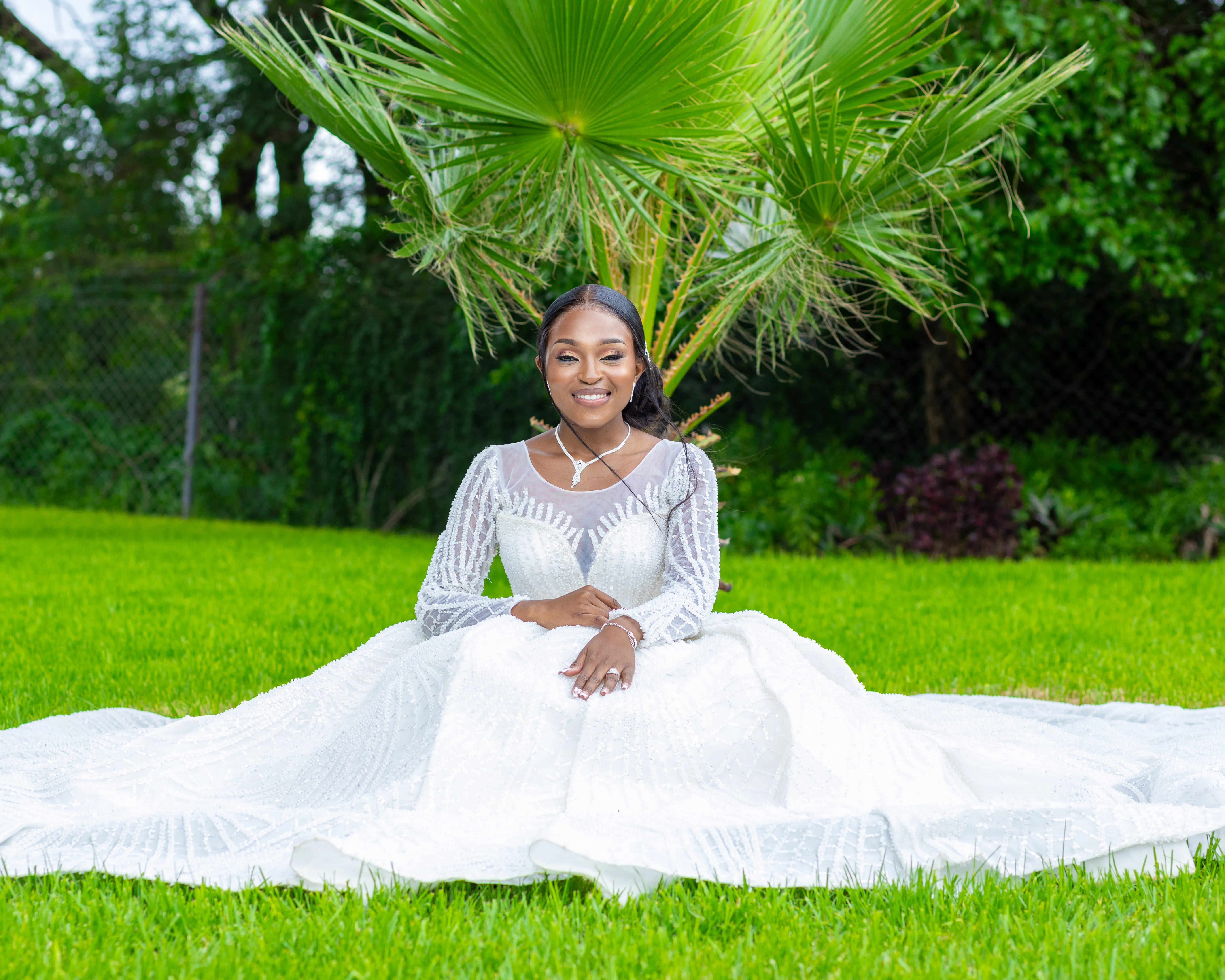 Smiling bride in a white lace wedding dress sitting on green grass with palm leaves in the background.