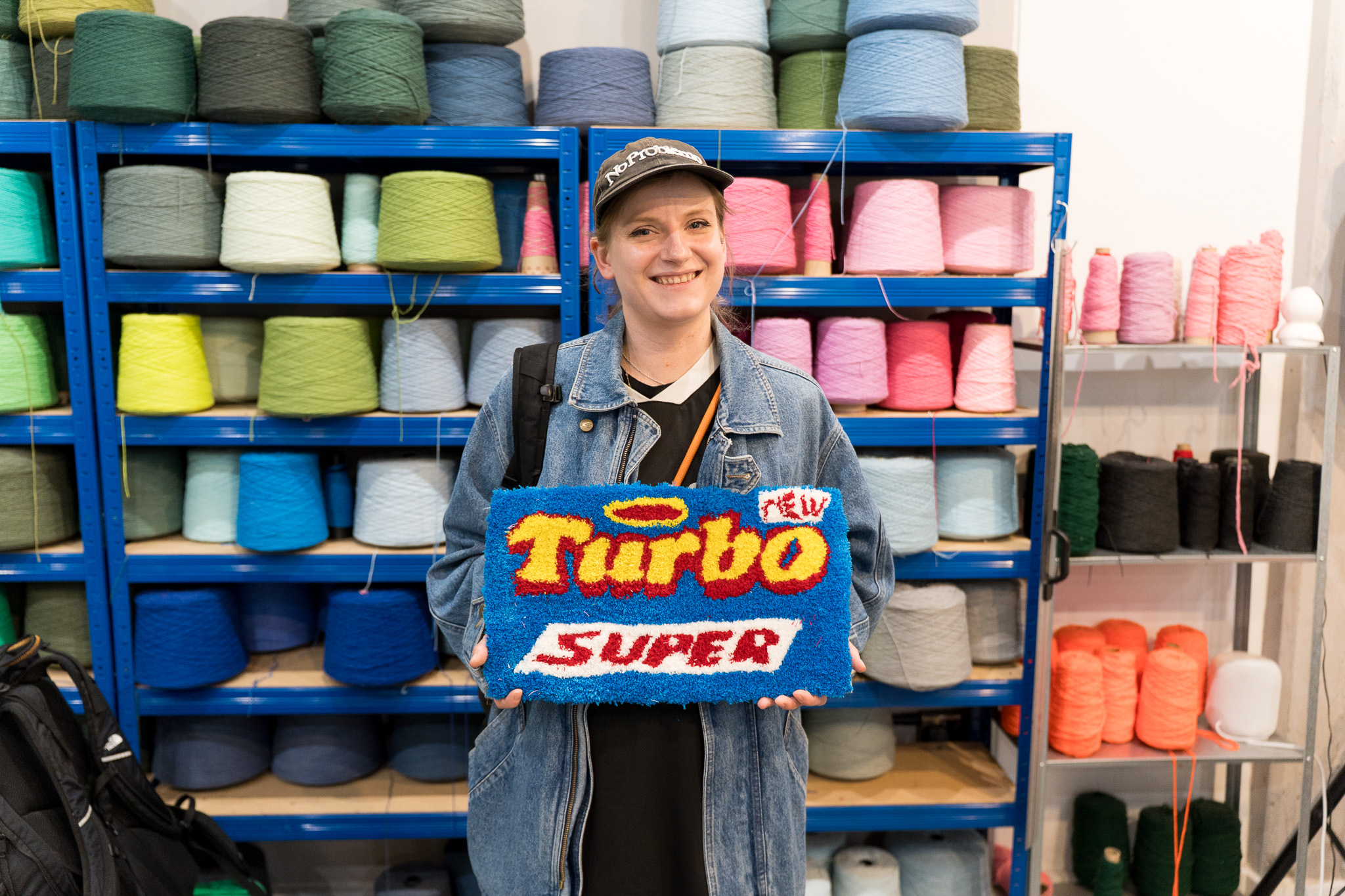 A smiling workshop participant holds up a tufted rug featuring the Turbo Super chewing gum logo in bold red, yellow and blue, standing in front of colourful yarn shelves at Cheeky Studio, Hackney Wick.