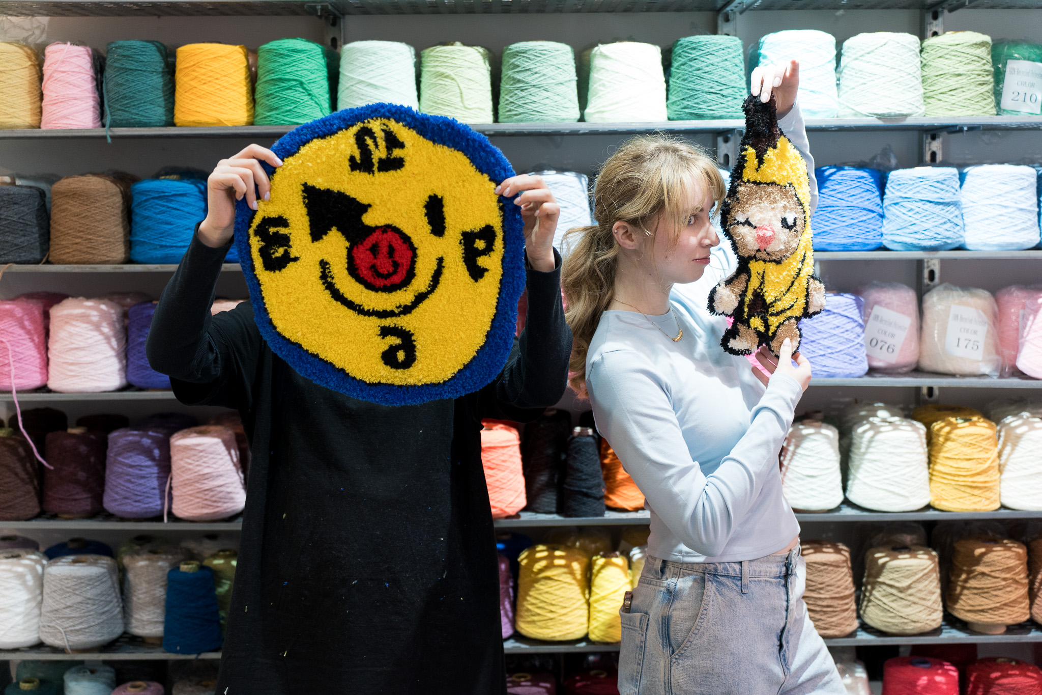 Two finished tufted rugs held up at Cheeky Studio — a yellow smiley face design and a cat figure — in front of shelves of coloured yarn cones