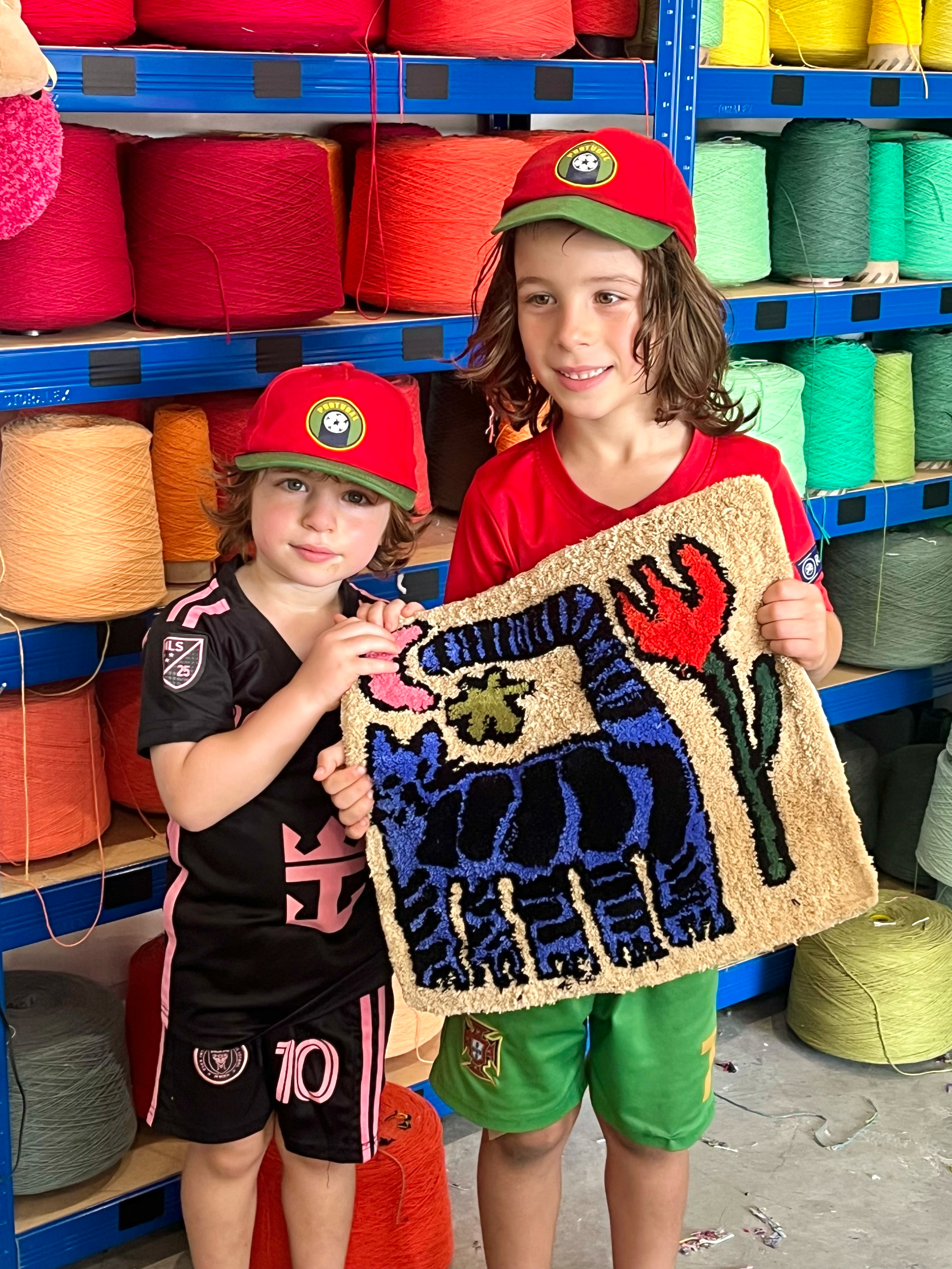 Two young children holding a finished tufted rug of a blue horse design at Cheeky Studio, in front of shelves of colourful yarn cones
