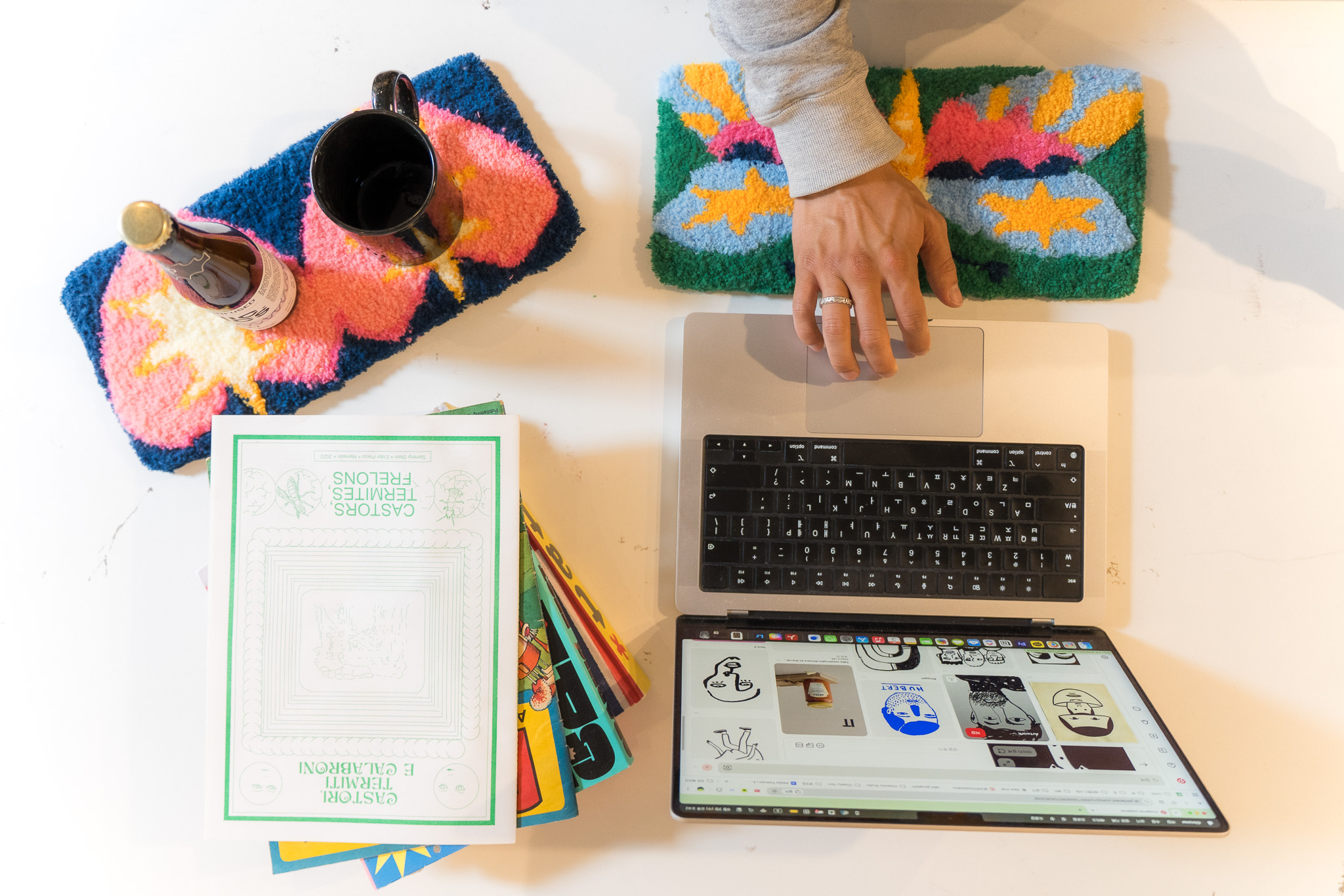 Close-up of a hand resting on a colourful tufted wrist rest while using a laptop, with a second tufted mat holding a mug and bottle nearby