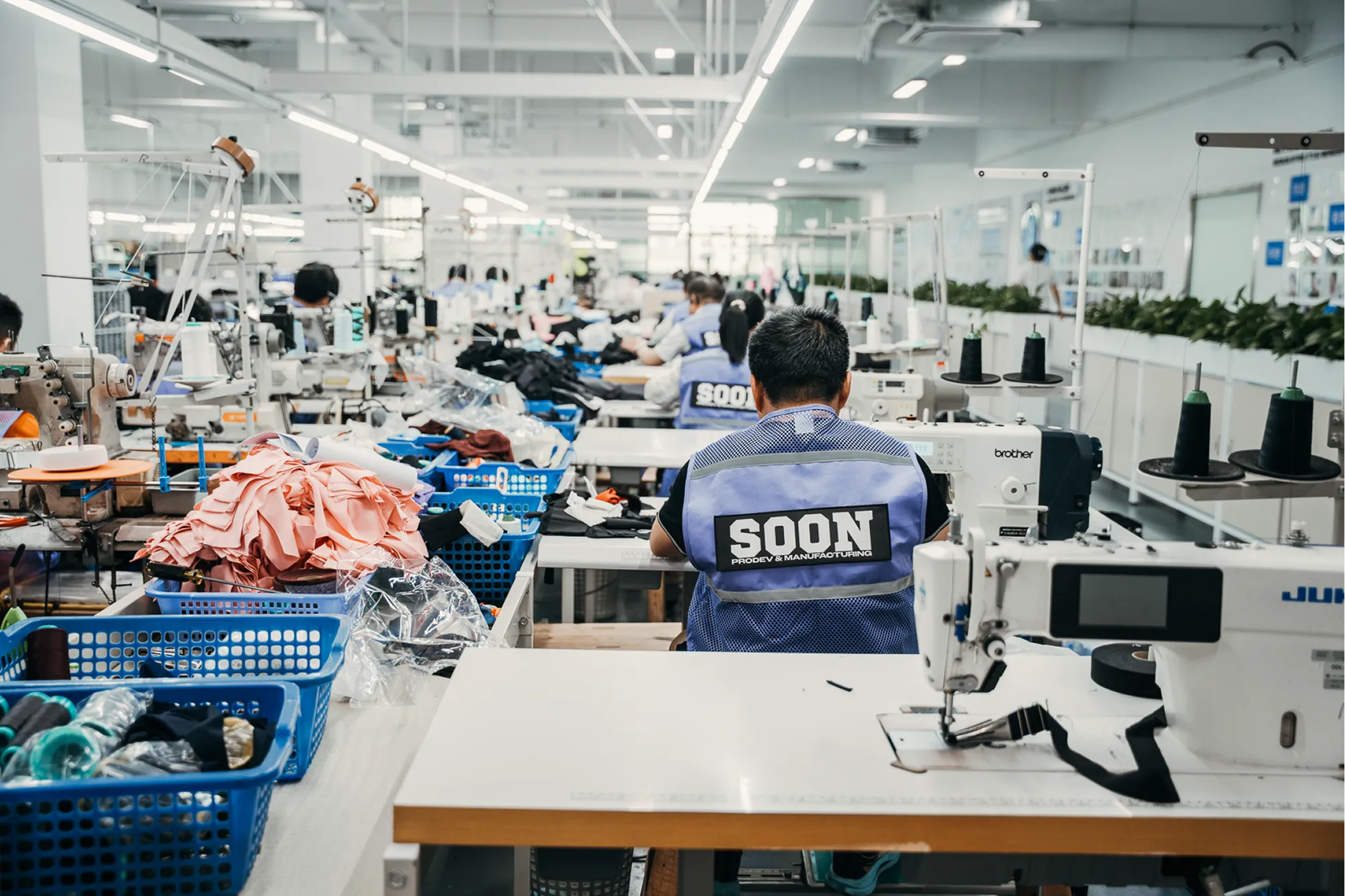 Factory workers wearing SOON vests operate sewing machines in a garment manufacturing facility.